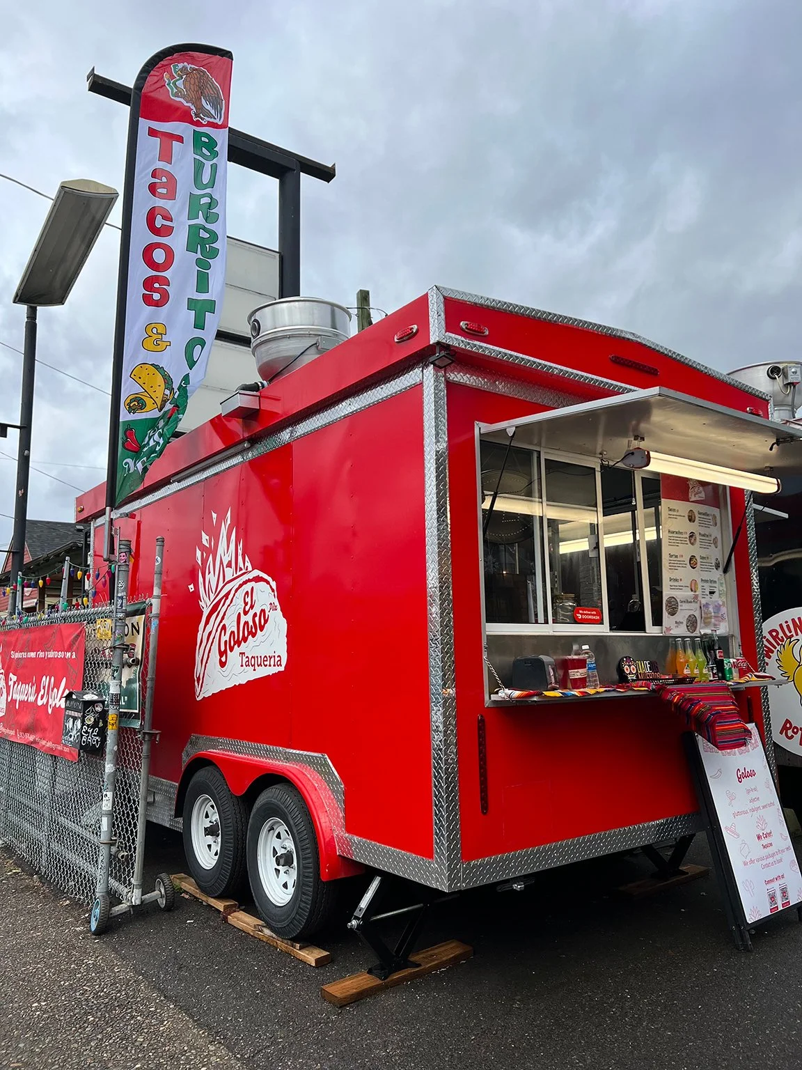 Red taco truck with a window display of menu items, a sign with the logo 'El Galoza Taqueria,' and a tall sign advertising burritos and tacos.