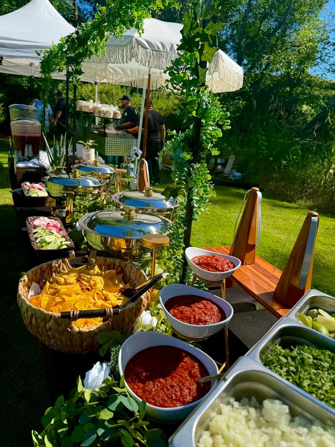 Buffet table outdoors with various bowls of salsa, chopped onions, cilantro, and tortilla chips, set in a grassy garden area with trees, umbrellas, and people serving food in the background.