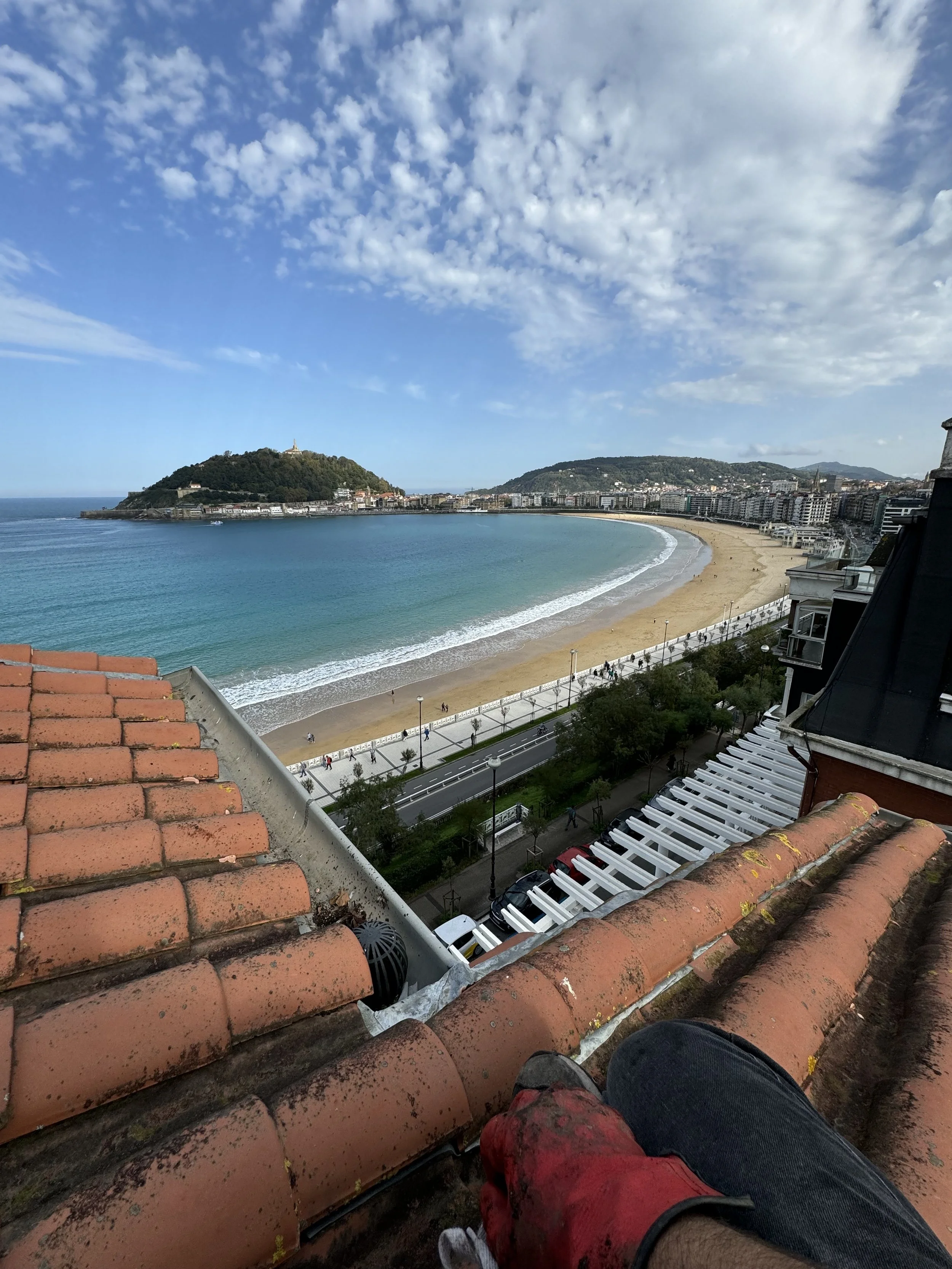 Vista panorámica de la playa de la Concha en Donostia con arena dorada, agua azul y una línea de edificios urbanos al fondo. Desde un edificio alto, se ve parte de un techo de tejas terracota en primer plano.