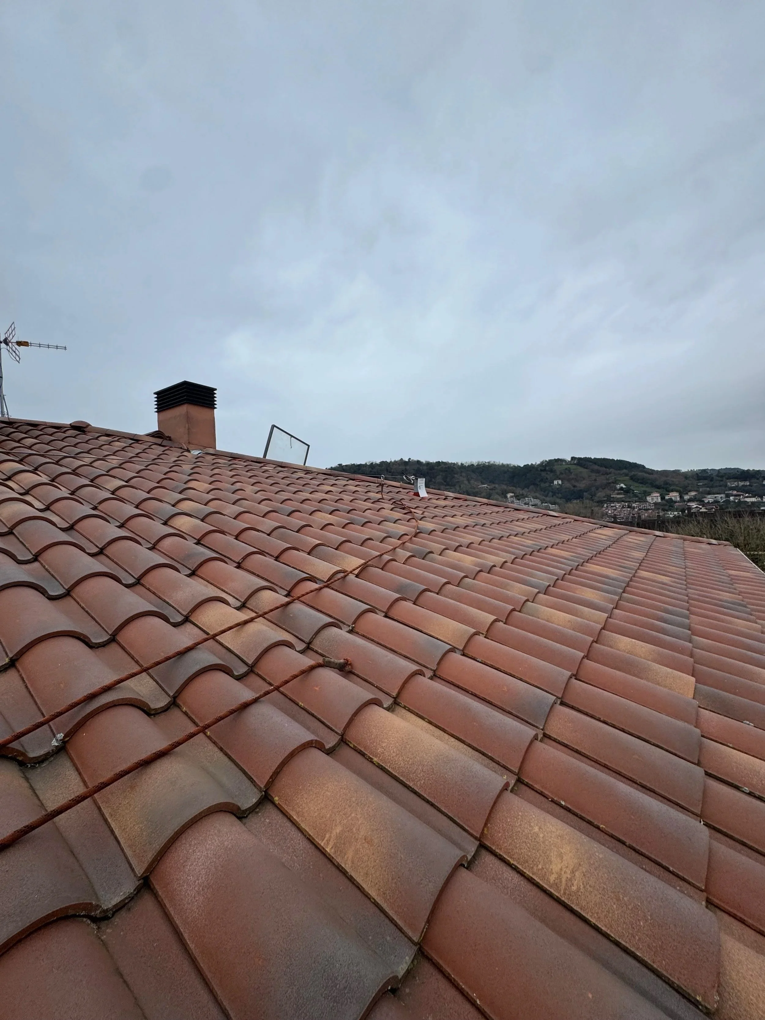 Tejado con tejas de color terracota y un cable con nudos en la superficie, con fondo de cielo nublado y colinas en la distancia.