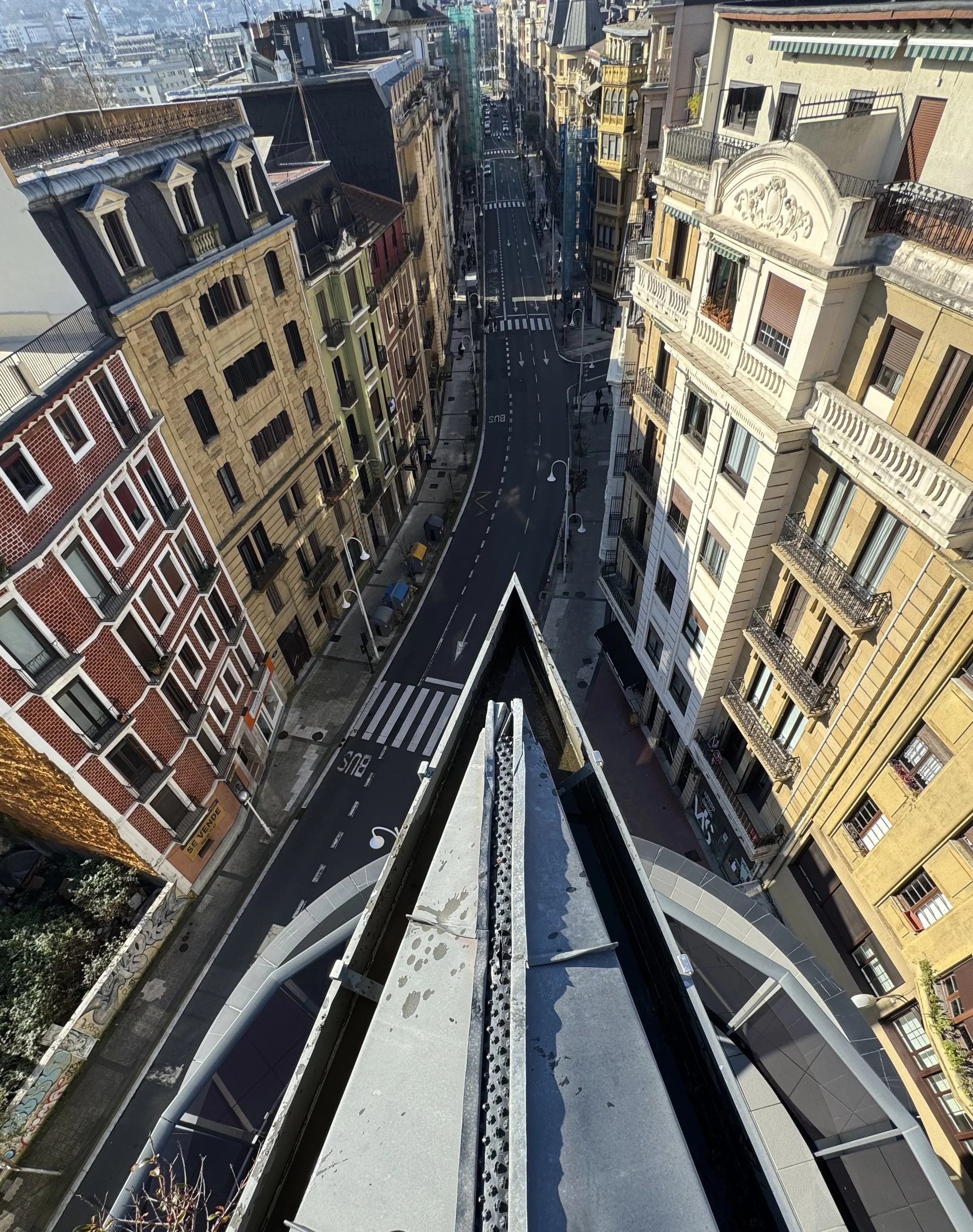 Vista de una calle en un área urbana desde una antena, con edificios altos y aceras en ambos lados. La calle tiene líneas blancas, señales de tránsito y un cruce peatonal.
