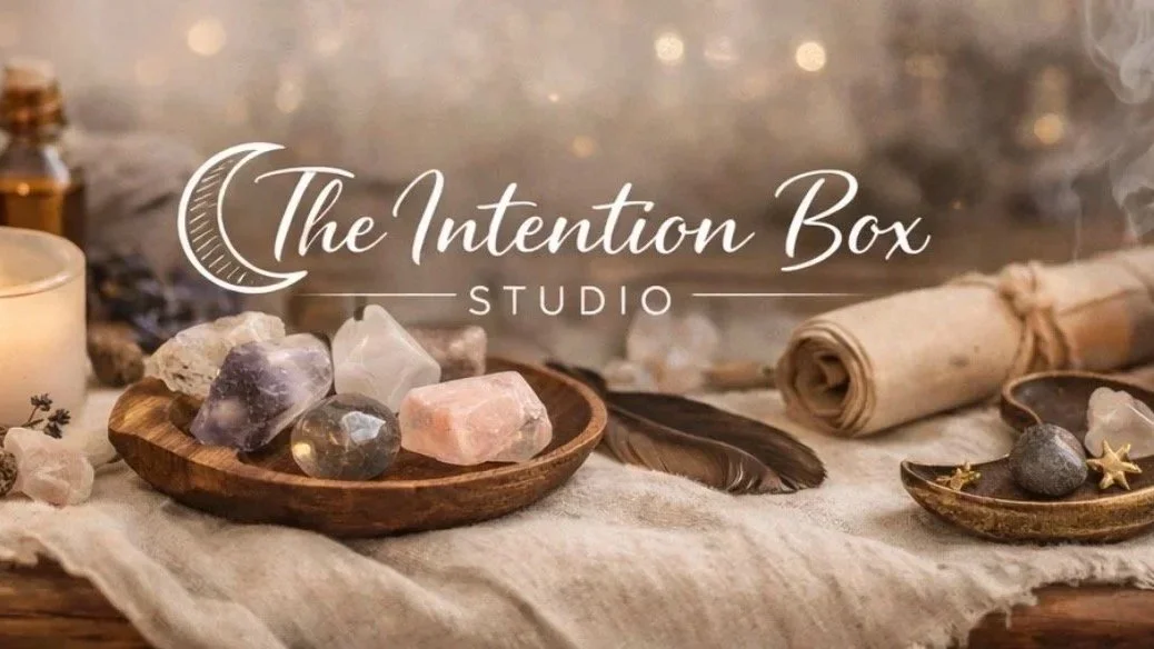 The logo of The Intention Box Studio with various crystals, a feather, a rolled scroll, and small decorative stones arranged on a soft cloth surface.