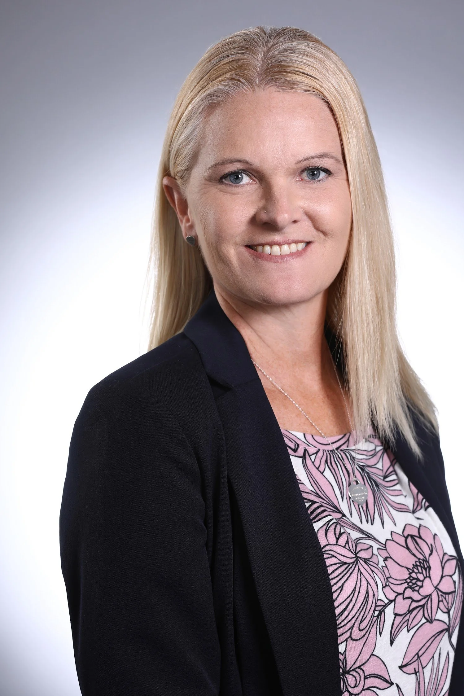 A professional woman with blonde hair, wearing a black blazer and a floral blouse, smiling at the camera against a neutral background.