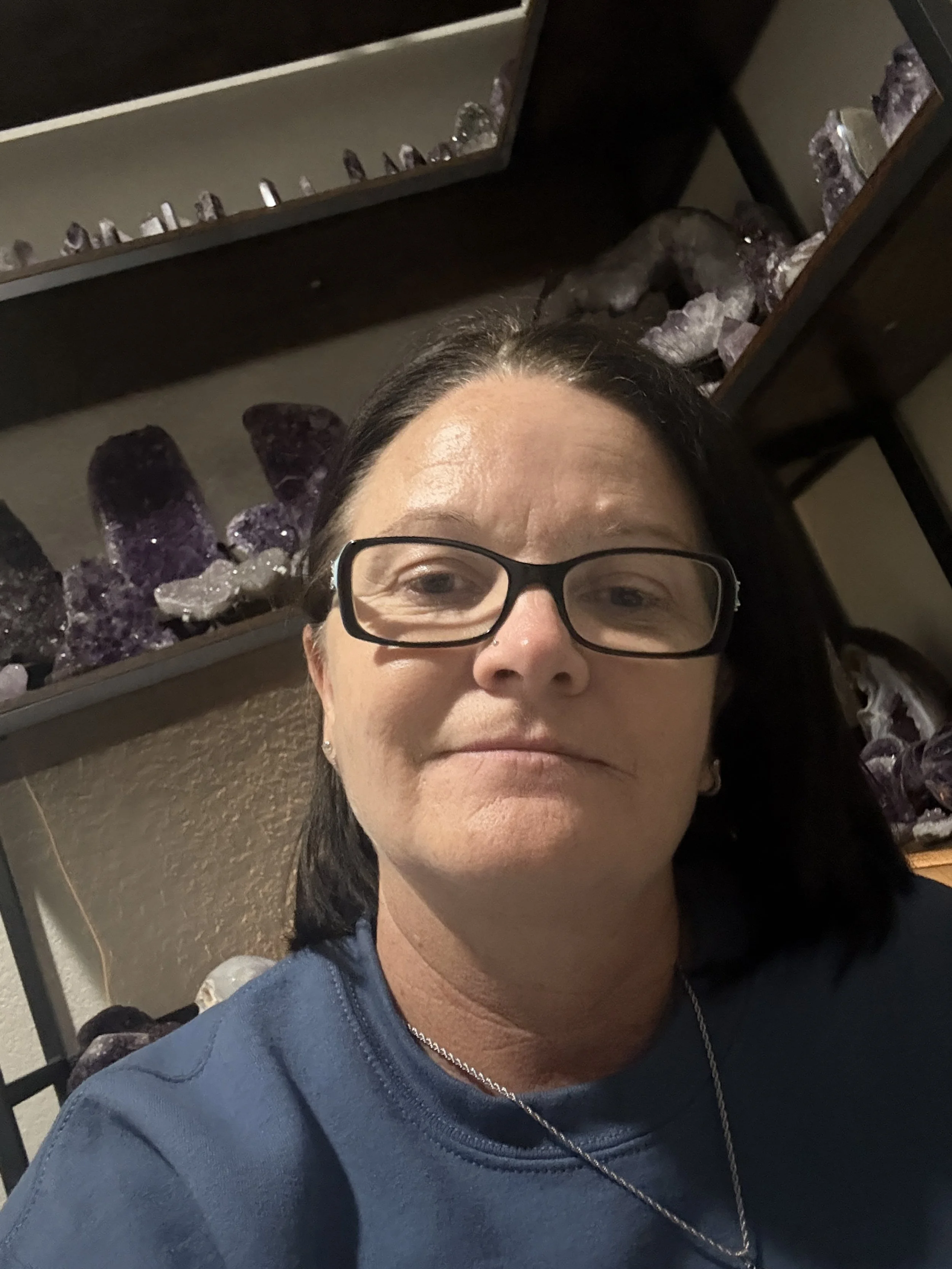 A woman with glasses taking a selfie in front of shelves with purple and white crystals.
