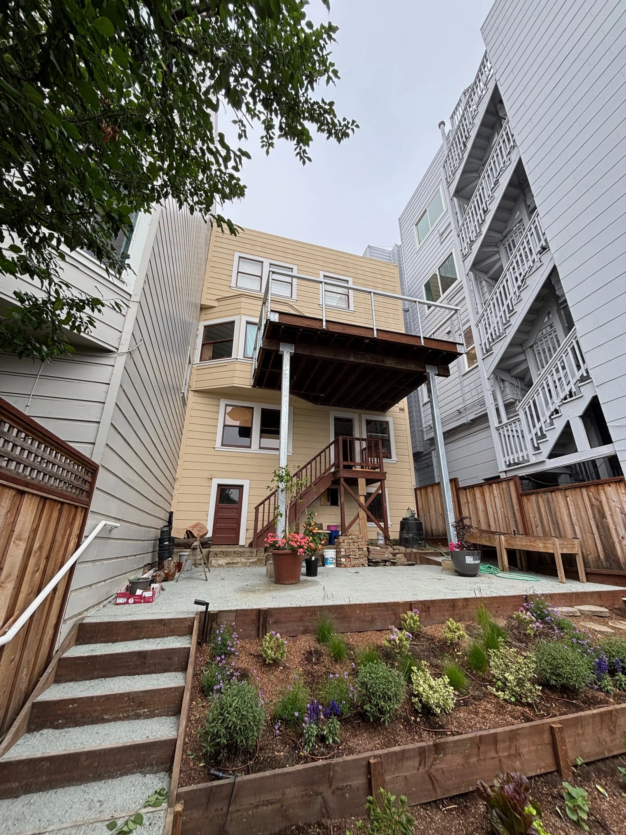Rear view of a multi-story house under renovation with open balcony, stairs, and garden in the foreground, surrounded by neighboring buildings.