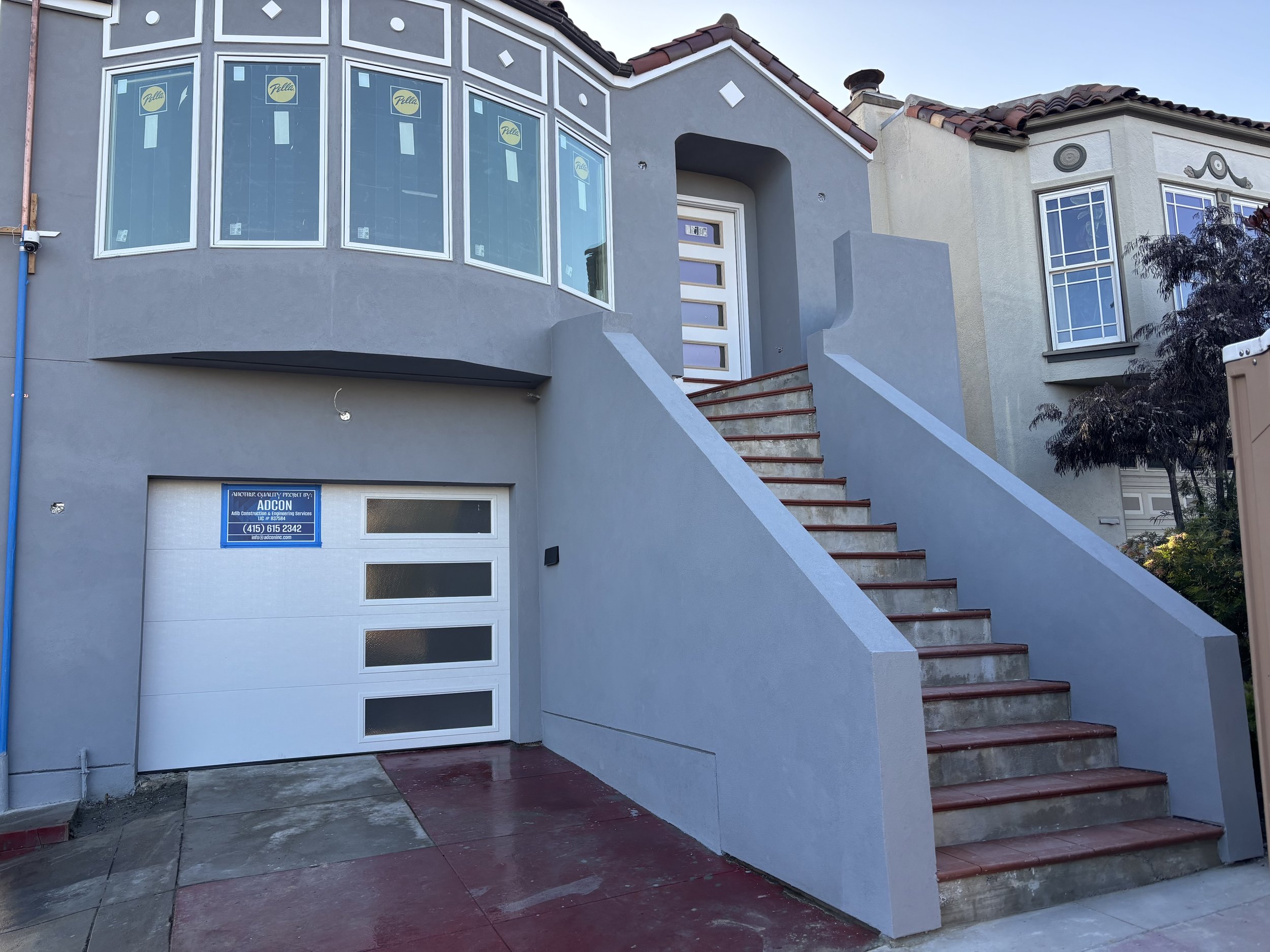 Front view of a modern house with a staircase leading up to the entrance, featuring a white garage door and large windows with some stickers, and a small window with bars next to the staircase, with a tree visible on the right side.