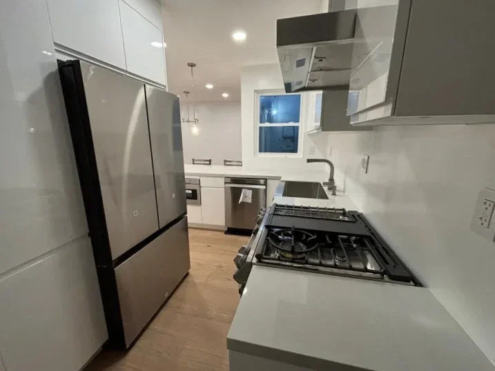 Modern kitchen with stainless steel refrigerator, white cabinets, and a gas stove with a white countertop. A window shows it is evening outside.
