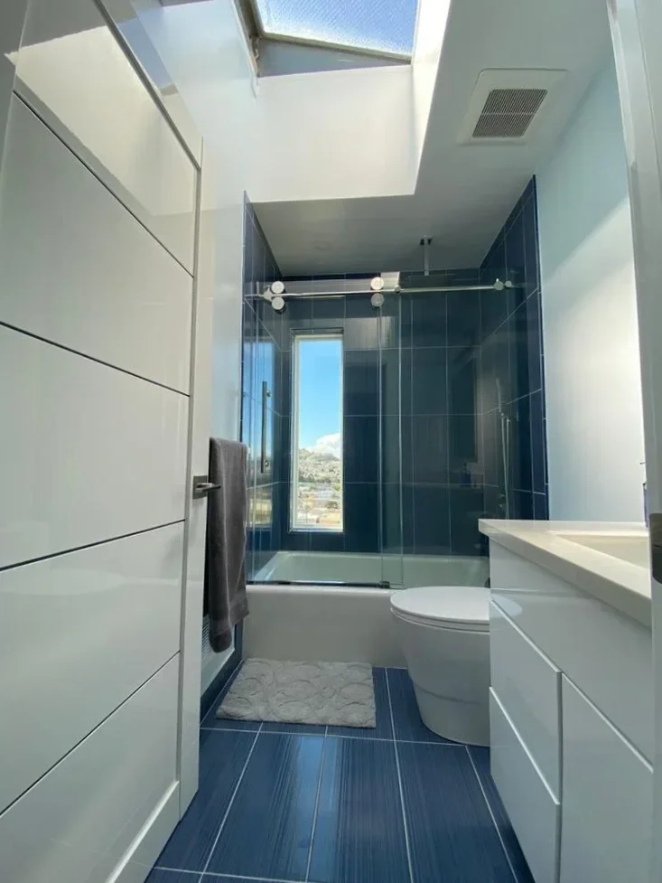 Modern bathroom with large window, blue tiled shower area with glass door, white vanity with sink, toilet, gray towel hanging, beige bath mat, and skylight ceiling.