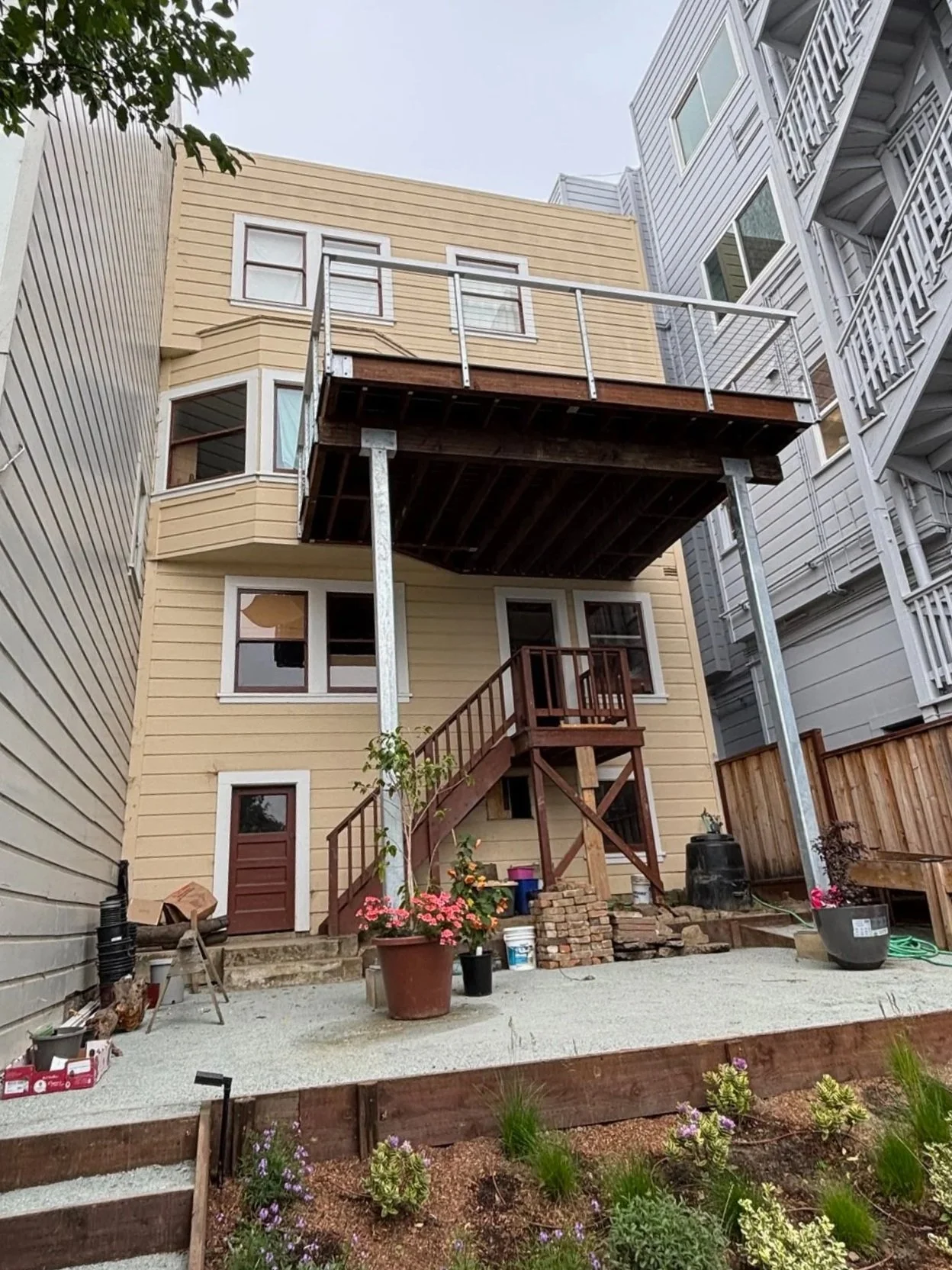 Back of a yellow house with a newly constructed wooden balcony on the second floor. There are planting pots and gardening supplies on the ground. The house is situated between other multi-story homes.