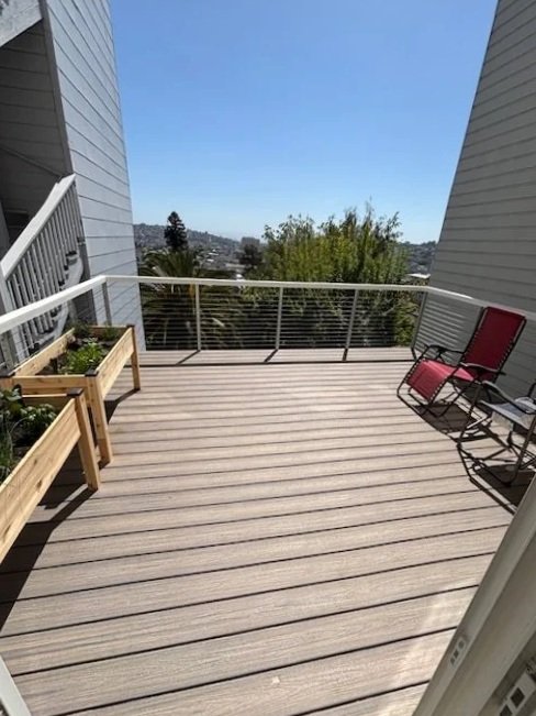Empty balcony with wooden deck, two red chairs, and potted plants, overlooking trees and a cityscape under a clear blue sky.