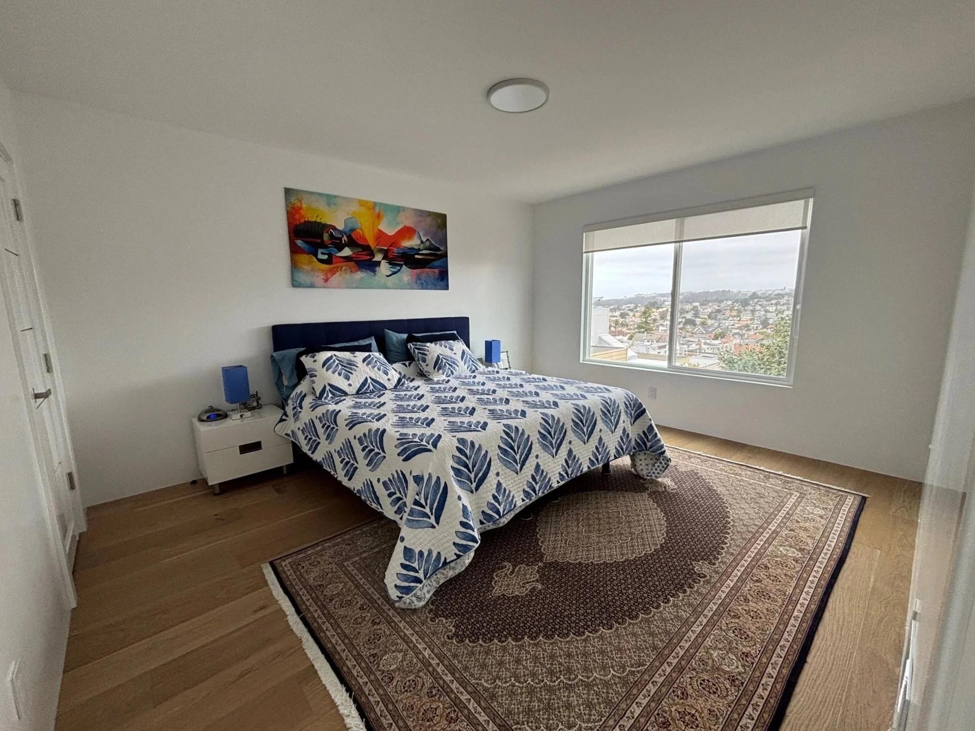 A bedroom with a bed, blue and white patterned bedding, a nightstand with a blue lamp, a window showing a cityscape, and a colorful abstract painting on the wall.