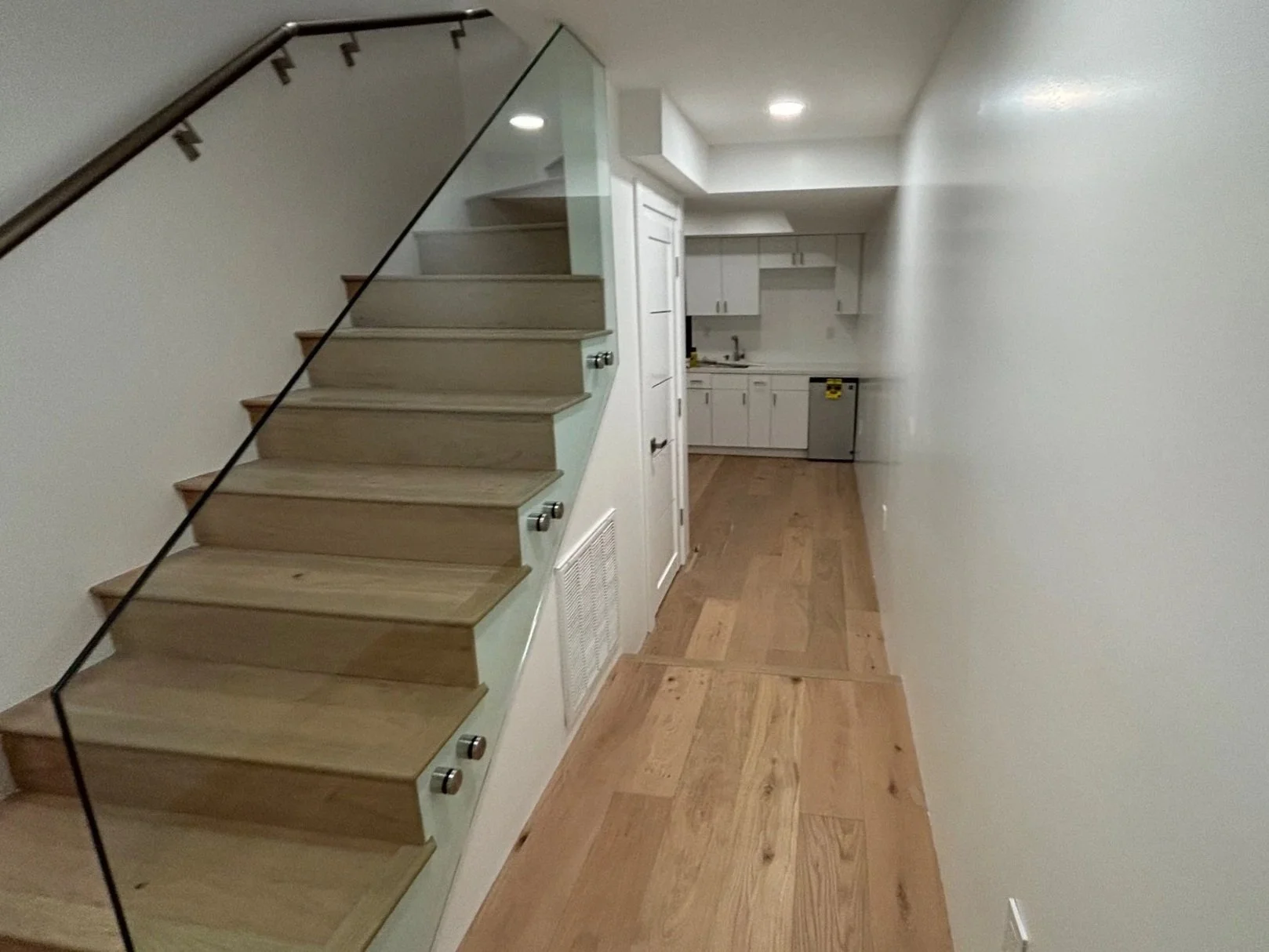 Interior view of a hallway with wooden flooring, a staircase on the left with glass railing, leading to an upper floor, and a kitchen in the background with white cabinets and wooden countertops.