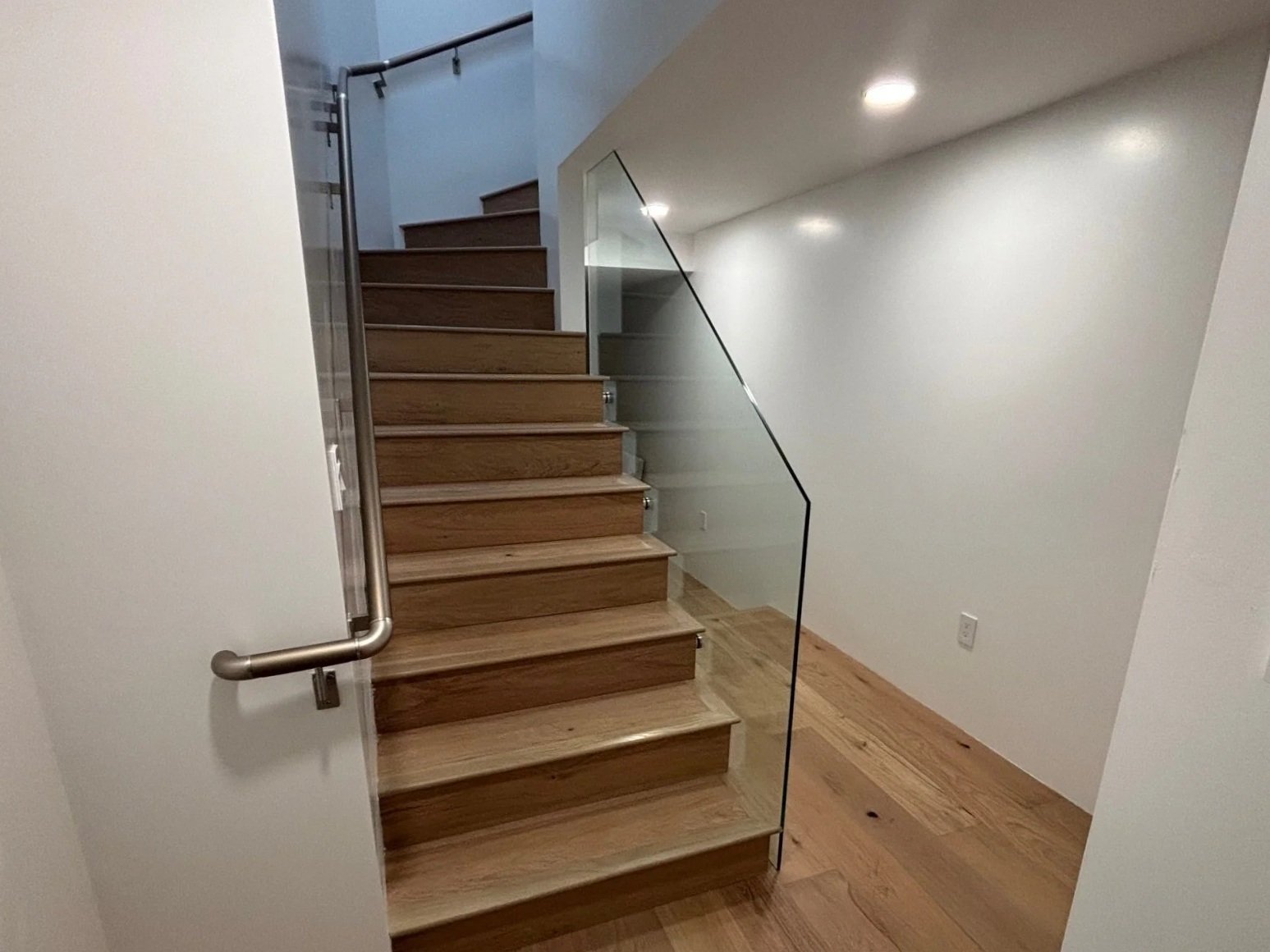 Wooden staircase with a glass railing and metal handrail, leading to an upper floor in a modern interior with white walls and hardwood flooring.