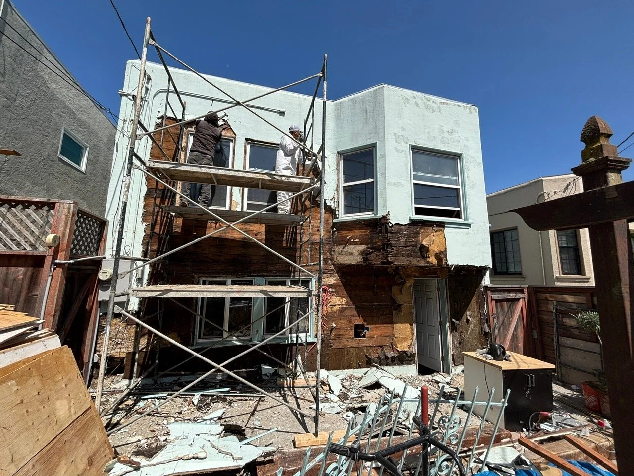 Construction workers renovating the exterior of a two-story house, with scaffolding and damaged wood, under a clear blue sky.