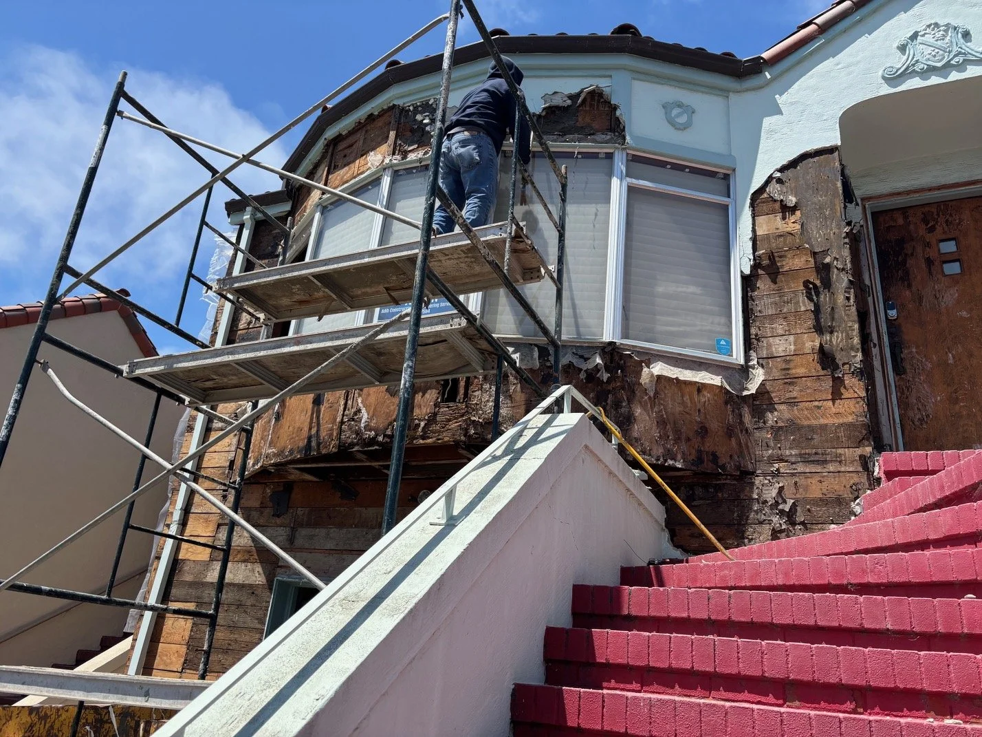 Person working on scaffolding outside a house, repairing or renovating the exterior wall which has exposed wood and damaged siding. The house has a large window and a decorative emblem above it, with stairs painted red leading up to the entrance.
