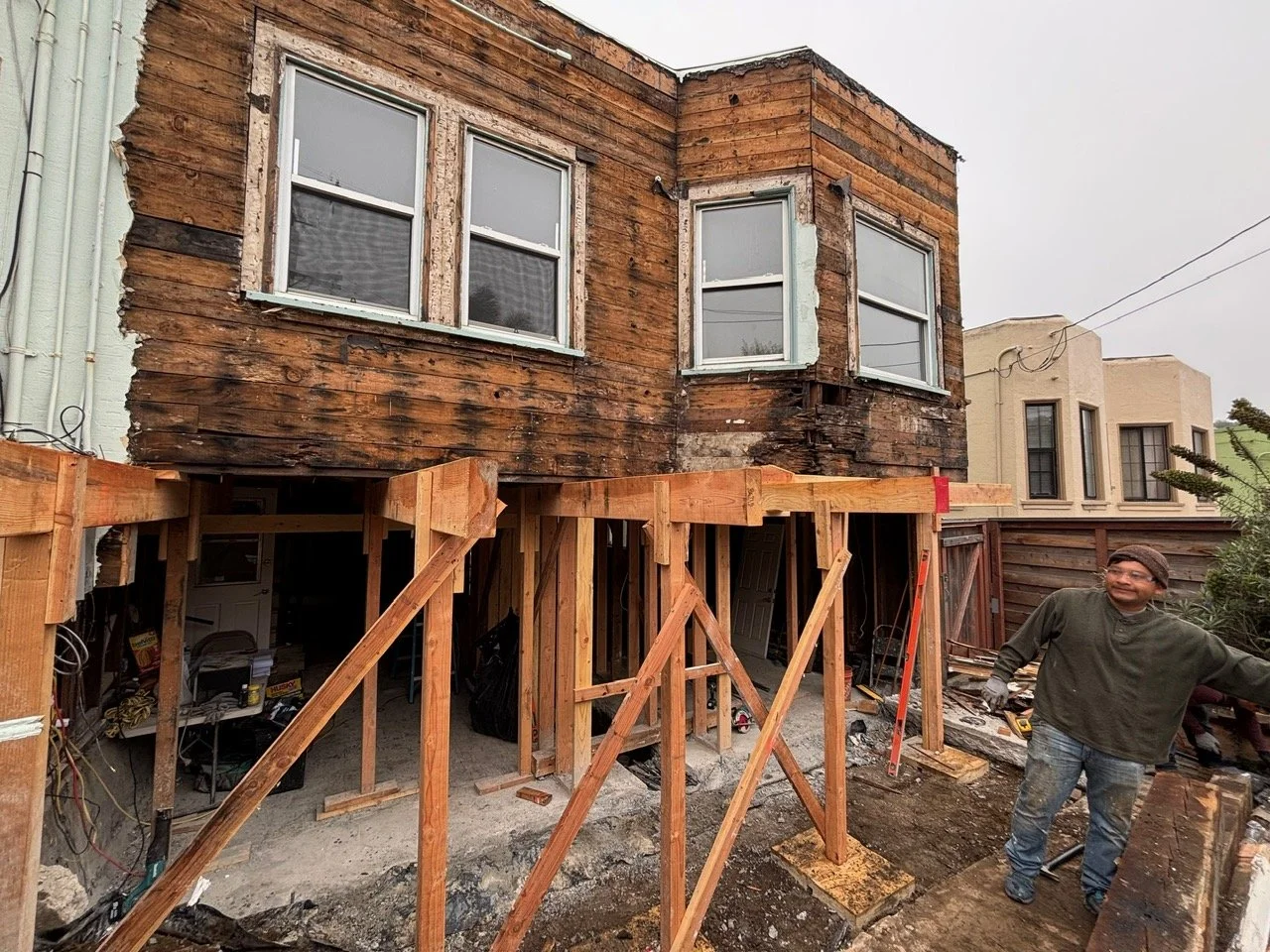 A house undergoing renovation, showing the exterior with exposed weathered wood siding on the upper level and newly installed wooden framing on the lower level, with a man standing to the right in a green shirt and jeans, smiling.