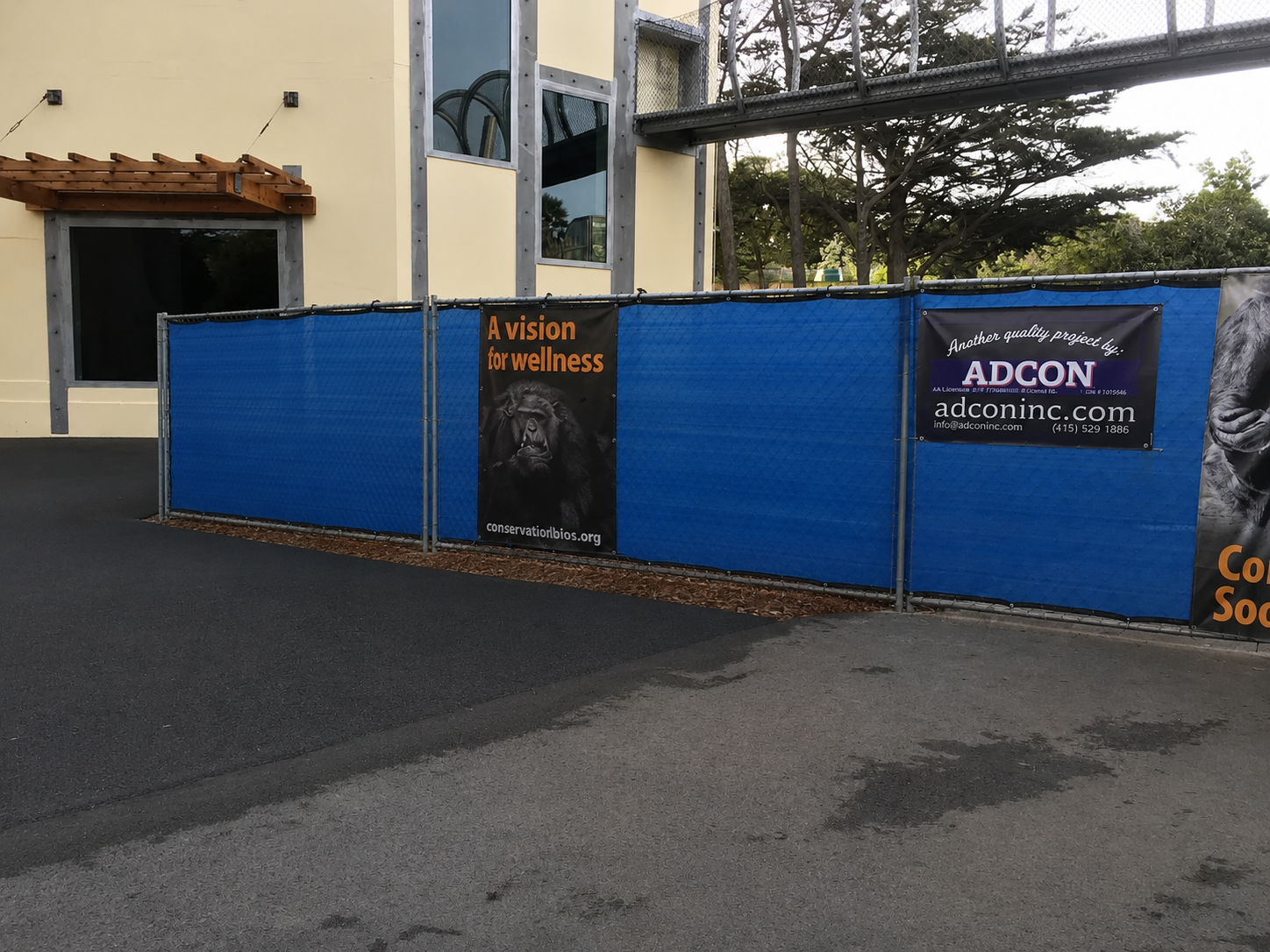 Blue construction fence with banners, one featuring a gorilla and the text 'A vision for wellness' and the website 'conservationbio.org', another for ADCON with contact information and the slogan 'Another quality project by ADCON', in front of a beig
