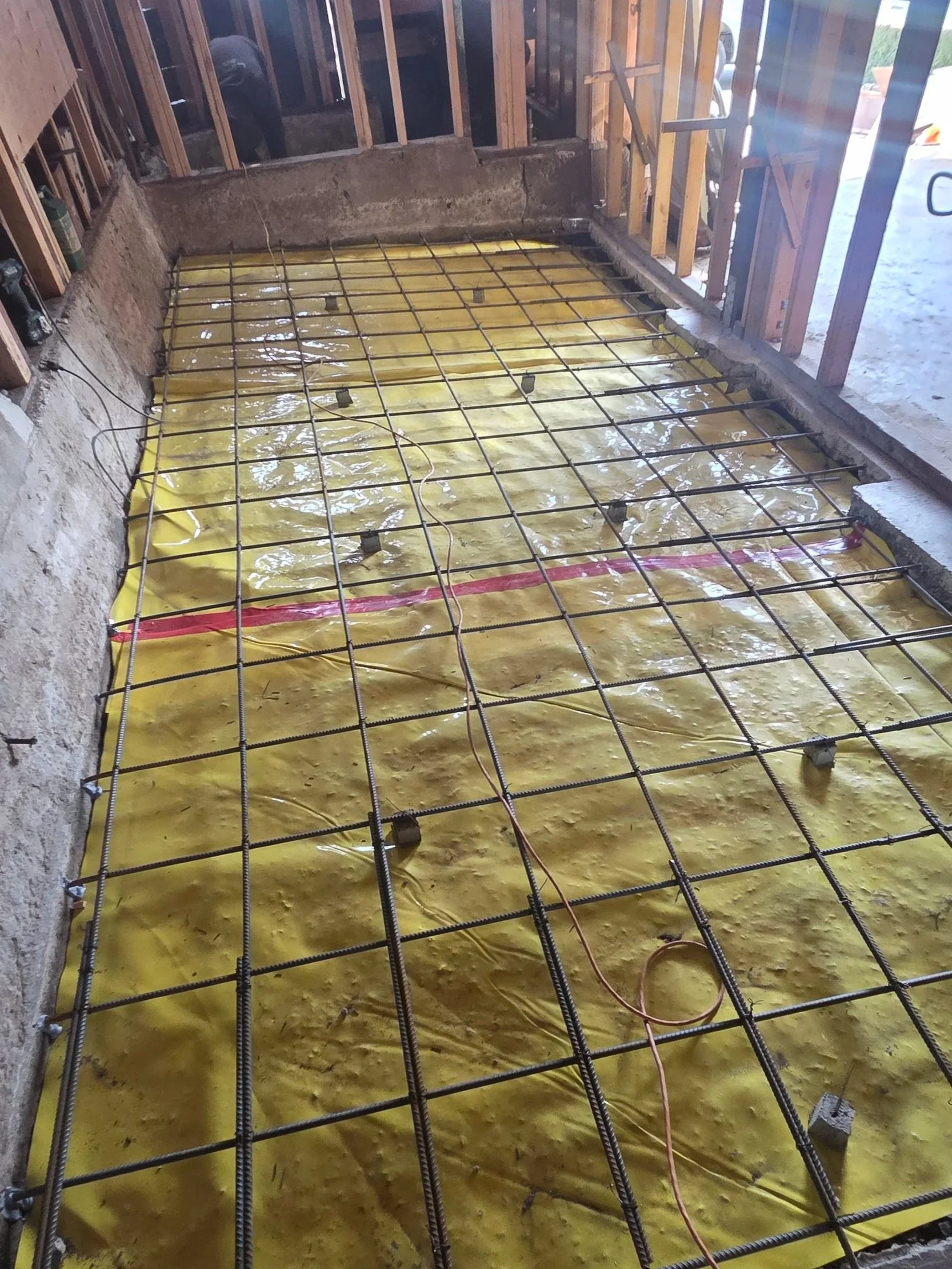 Construction site with a concrete slab foundation, yellow plastic vapor barrier, and steel rebar grid for reinforcing concrete, surrounded by unfinished wooden framing.