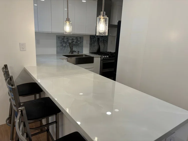 Modern kitchen with a white countertop island, three black bar stools, pendant lights, white cabinets, stainless steel sink, stove, and a gray backsplash with nature scene mural.