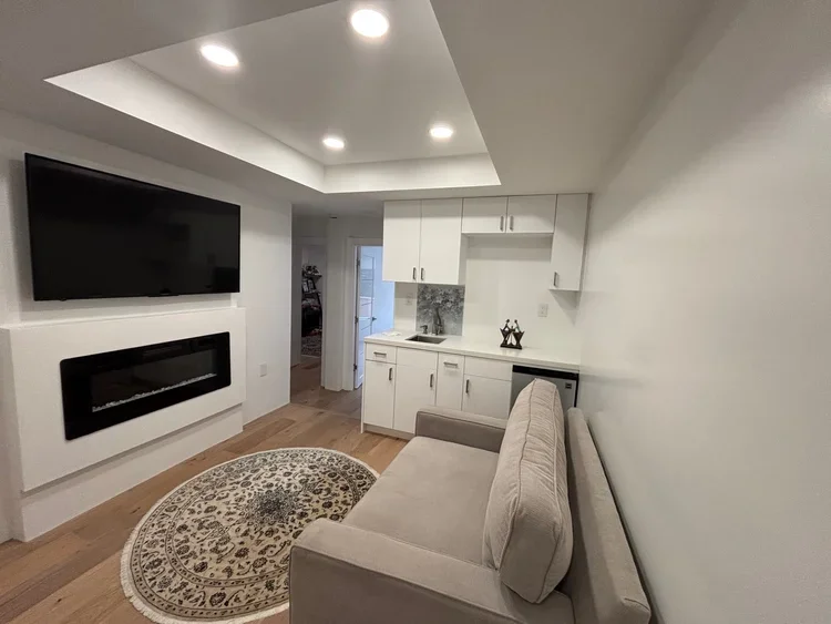 Living room with a wall-mounted flat-screen TV, electric fireplace, beige sofa, round patterned rug, and a small kitchen area with white cabinets and a marble backsplash.