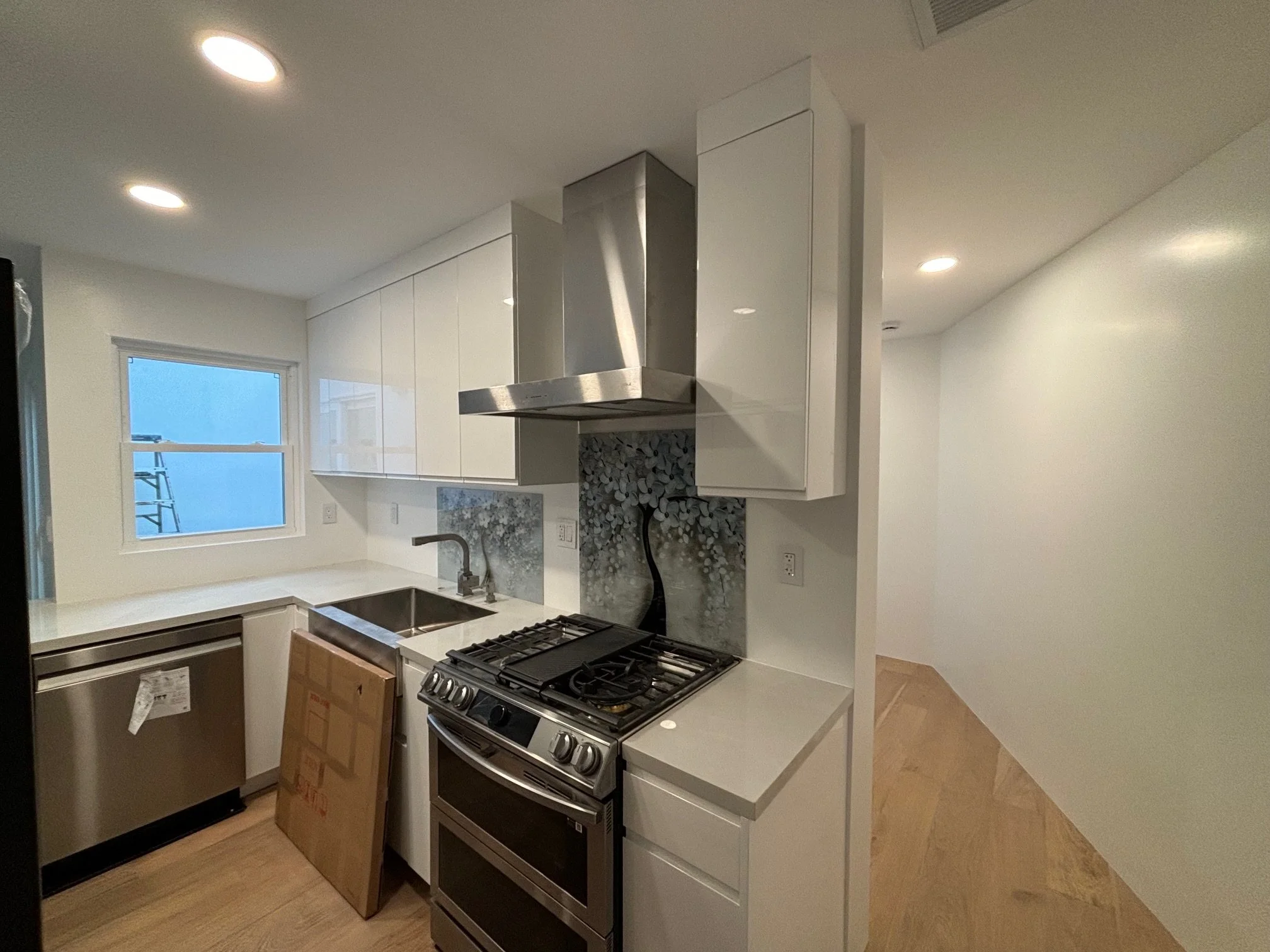 Modern kitchen with white cabinets, stainless steel appliances, wooden floor, window, and a small decorative backsplash behind the stove.