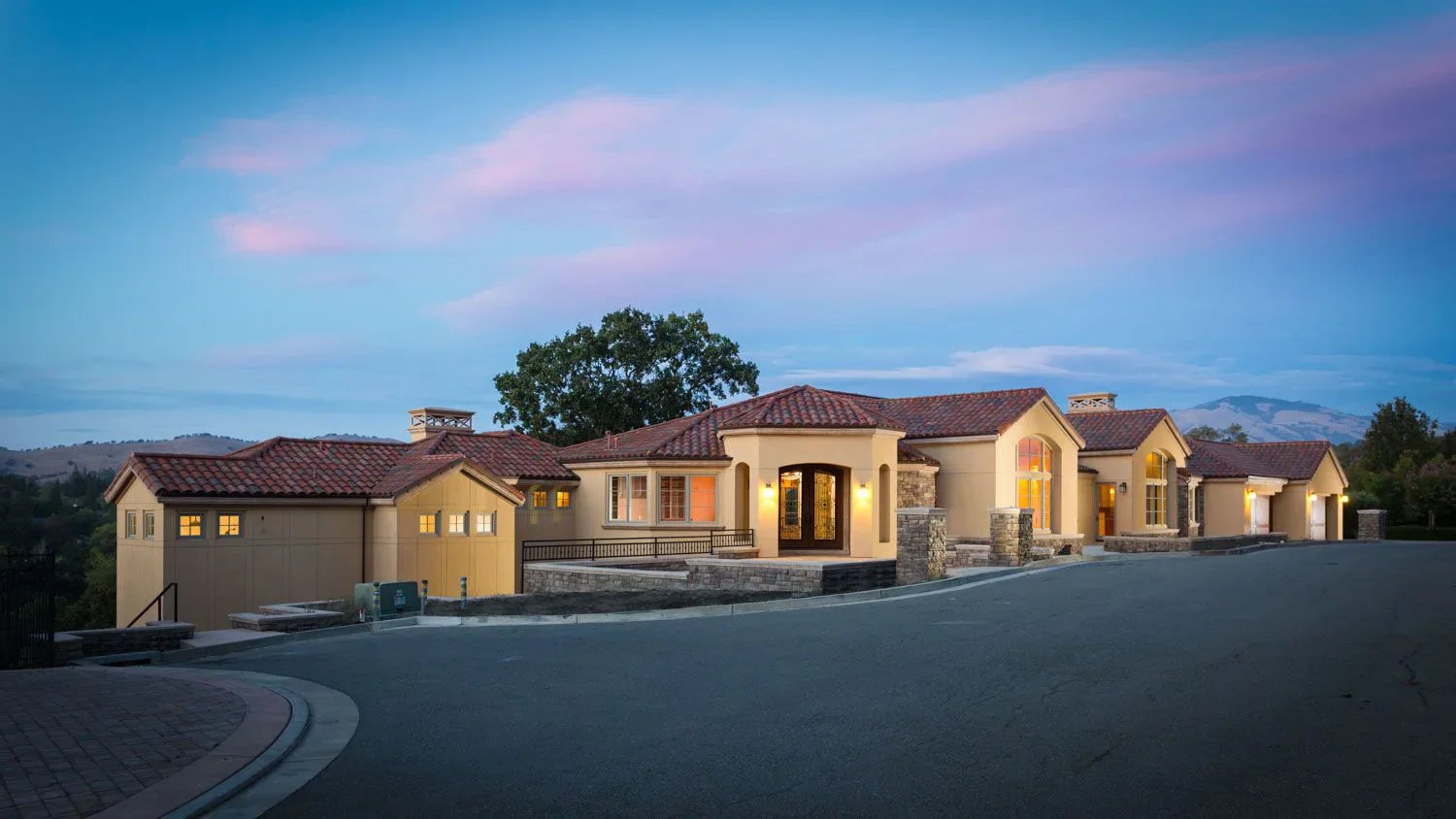 Large luxury house with a red-tiled roof, cream-colored walls, and stone accents, situated on a curved driveway at dusk with the sky transitioning from blue to pink.