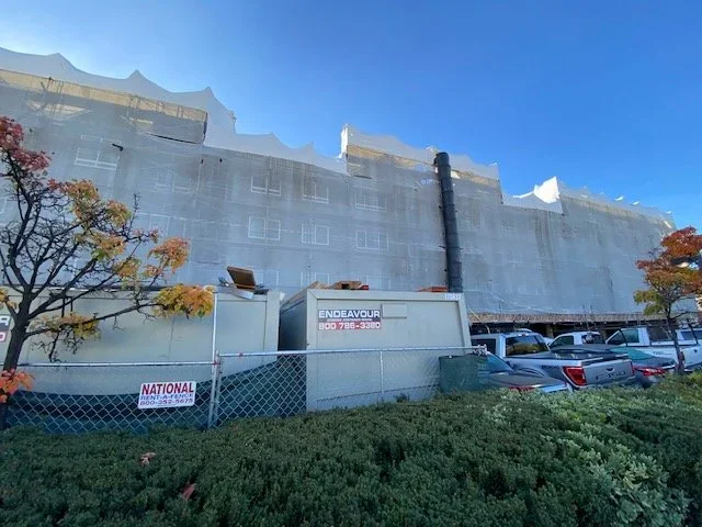 Building covered with protective mesh or netting, construction site with parked cars, trees with autumn foliage, and a clear blue sky.