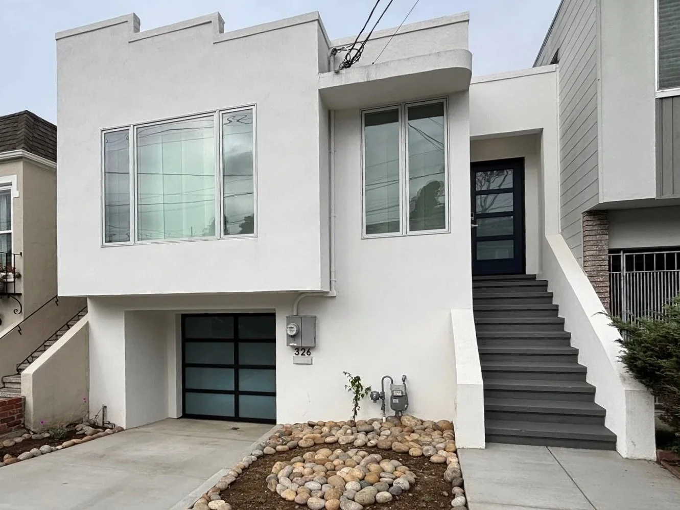 Front view of a modern white two-story house with a garage, stairs leading to the main entrance, large windows, and minimal landscaping with rocks.