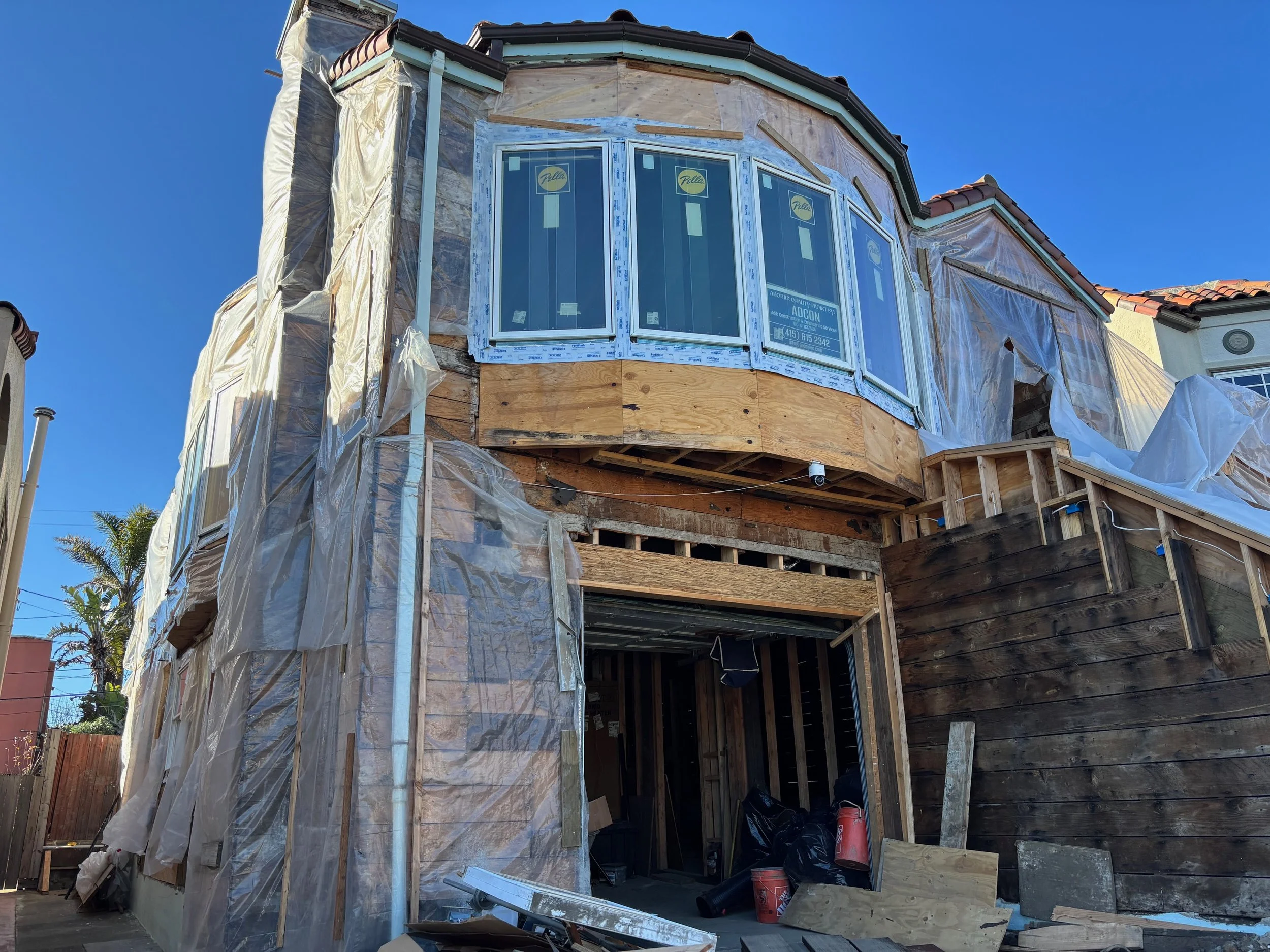 The exterior of a house under construction with scaffolding and exposed wooden framework, with a new bay window installed on the upper level and a partially built wall on the lower level, surrounded by construction materials and tools.