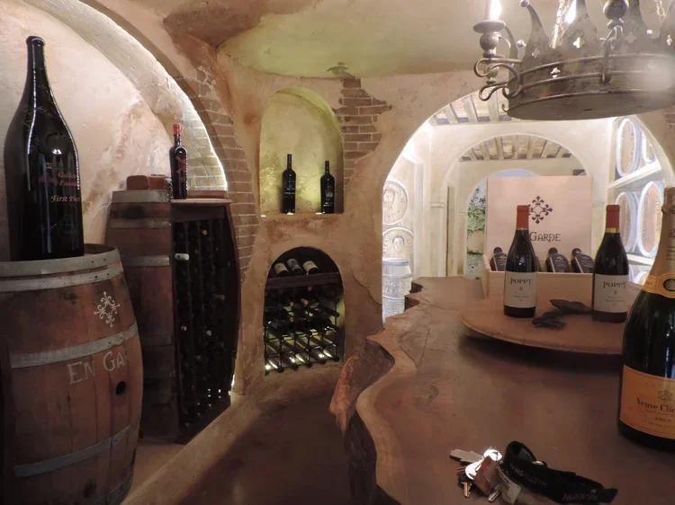 Wine bottles on a wooden wine barrel, wine rack with bottles, and a round wooden bar countertop with more wine bottles inside a rustic wine cellar with arched ceiling and brickwork.
