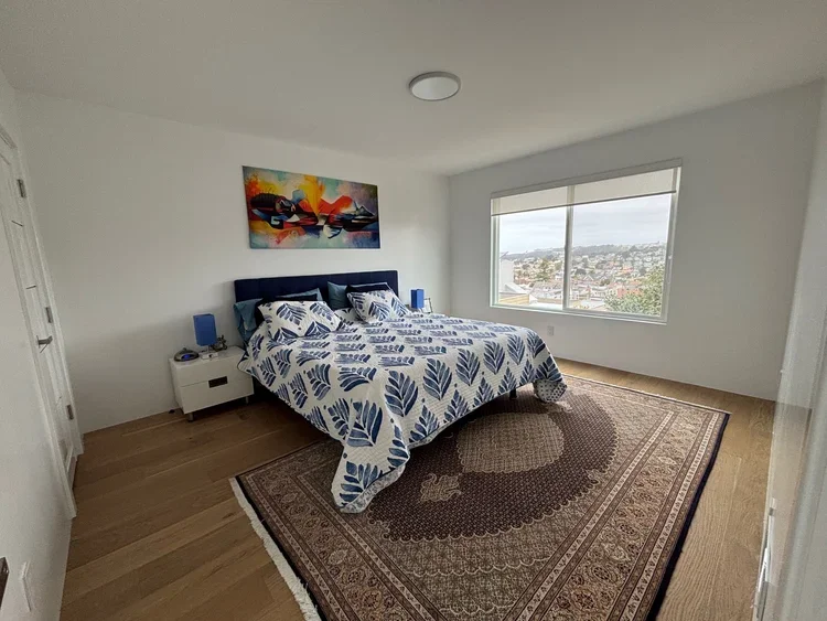 A bedroom with a large bed, blue headboard, and a patterned bedspread, two blue nightstands with lamps, a colorful painting above the bed, a large window displaying a cityscape, and a decorative area rug on wooden flooring.