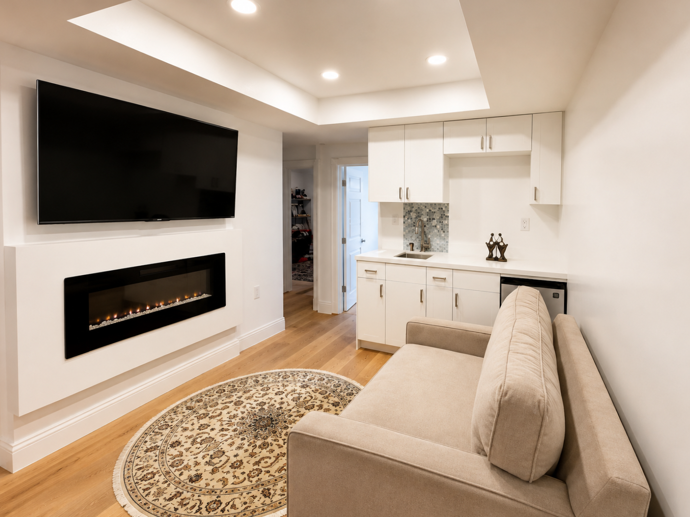 Living room with wall-mounted TV and fireplace, beige sofa, small kitchen area with white cabinets, small refrigerator, and decorative items, hardwood floor, and a round patterned rug.