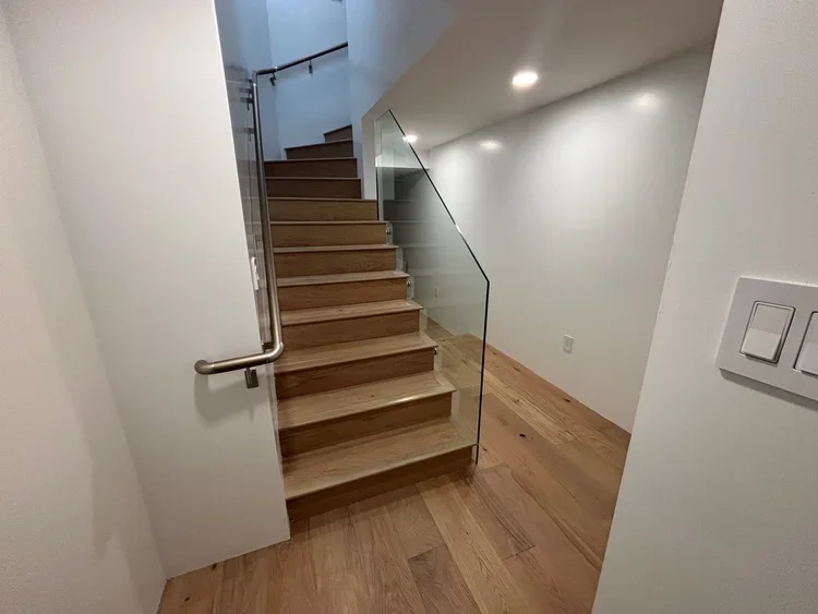 Wooden staircase with glass railing in a modern home, white walls, and wood flooring.
