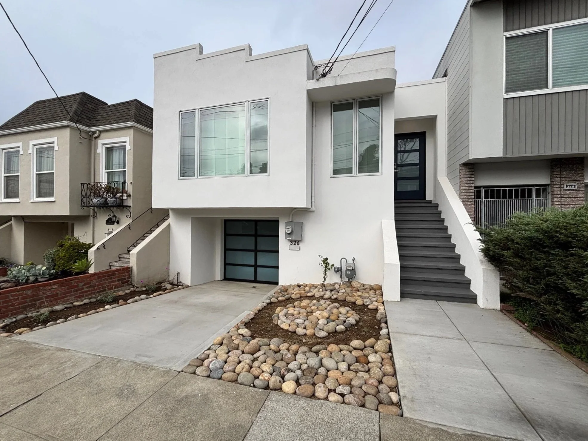 White modern two-story house with black door, glass garage door, staircase on right side, and small landscaped yard with rocks.