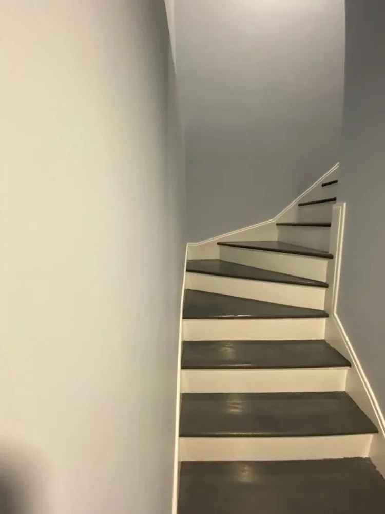 Interior view of a staircase with dark treads and white risers, ascending around a corner, with a blue wall and white baseboard.
