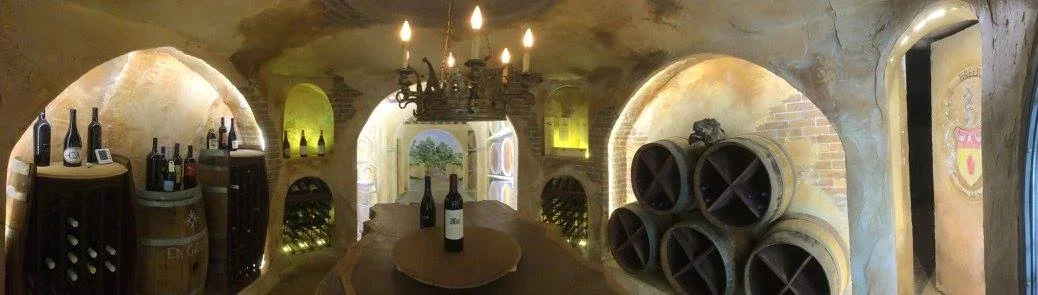 Wine bottles and barrels in a wine cellar with medieval-style stone walls and arched openings.