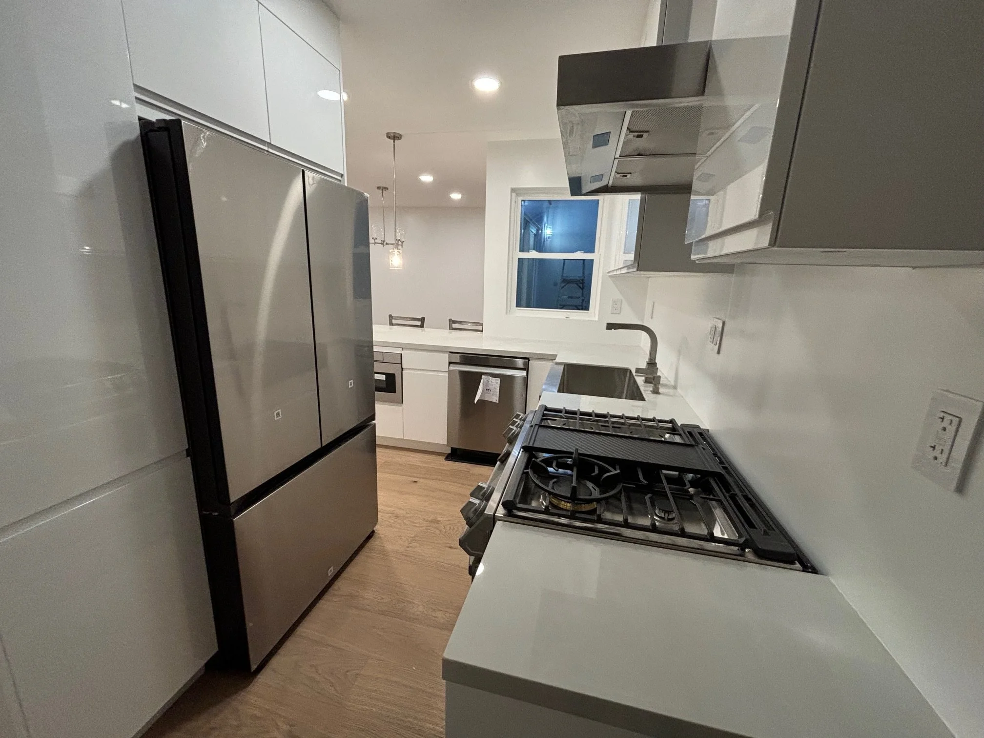 Modern kitchen with stainless steel refrigerator, white cabinetry, white countertops, gas stove, stainless steel sink, dishwasher, and a window with a view outside at night.