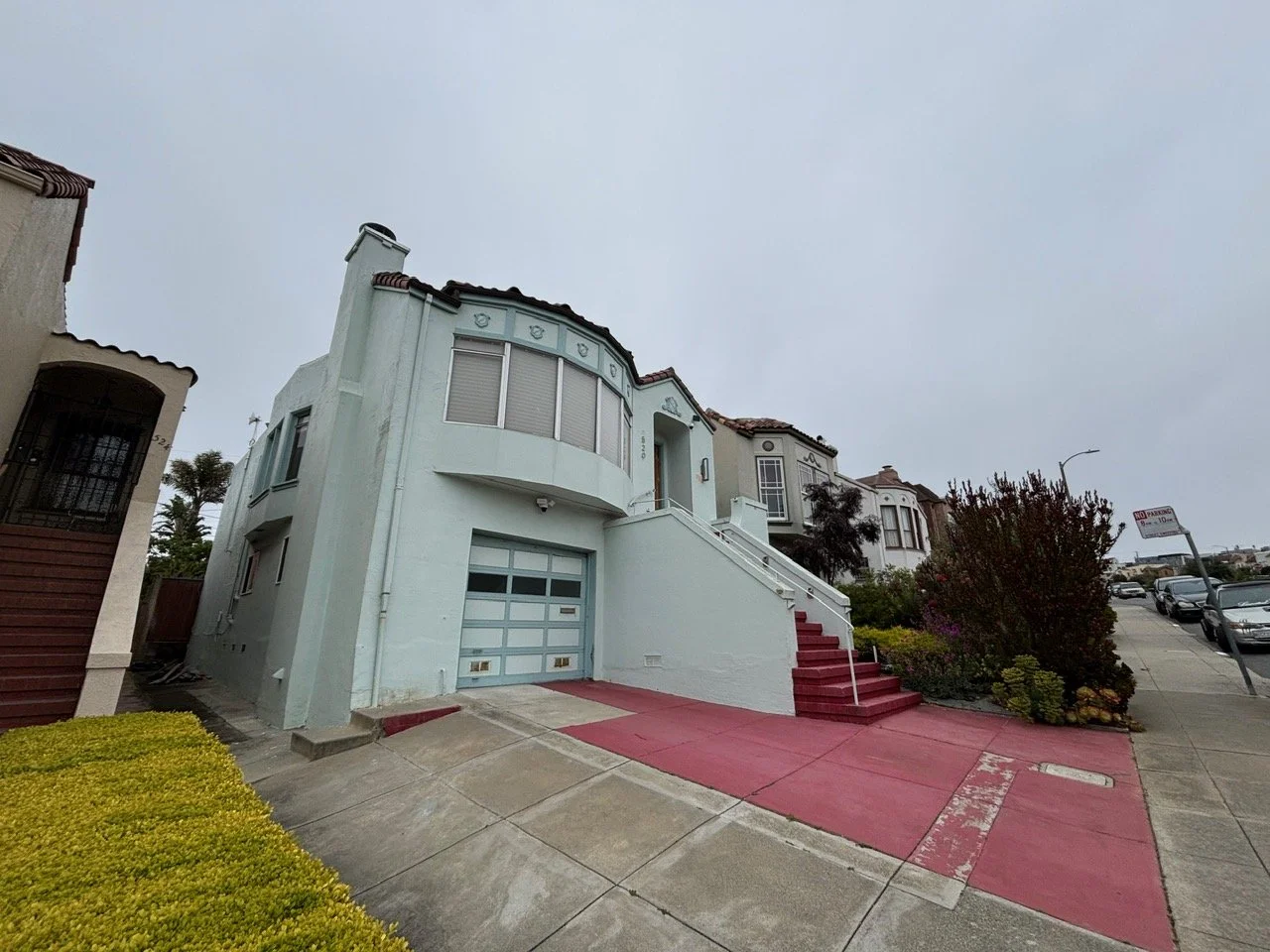 A pale blue multi-story house on a sloped street with a red driveway, stairs, and a garage with a glass paneled door, neighboring houses, and parked cars.