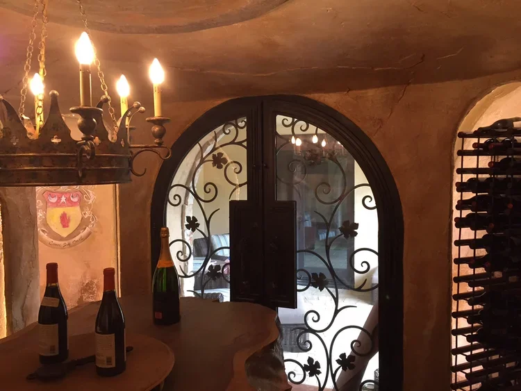Interior of a wine cellar with a chandelier, wine bottles on a curved wooden counter, a decorative wrought iron door, a cellar wall with a shield coat of arms, and a wine rack.