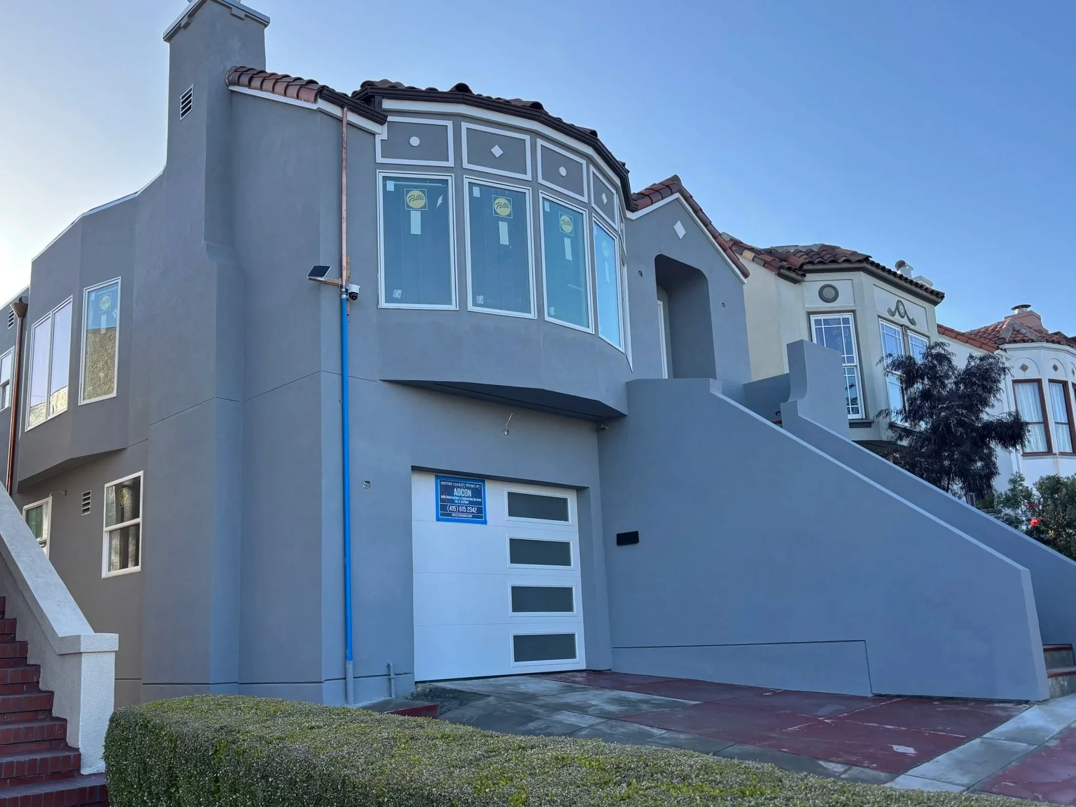 Front view of a multi-story house with gray exterior walls, red-tiled roof, several windows, and a staircase leading up to the entrance. Clear blue sky in the background.