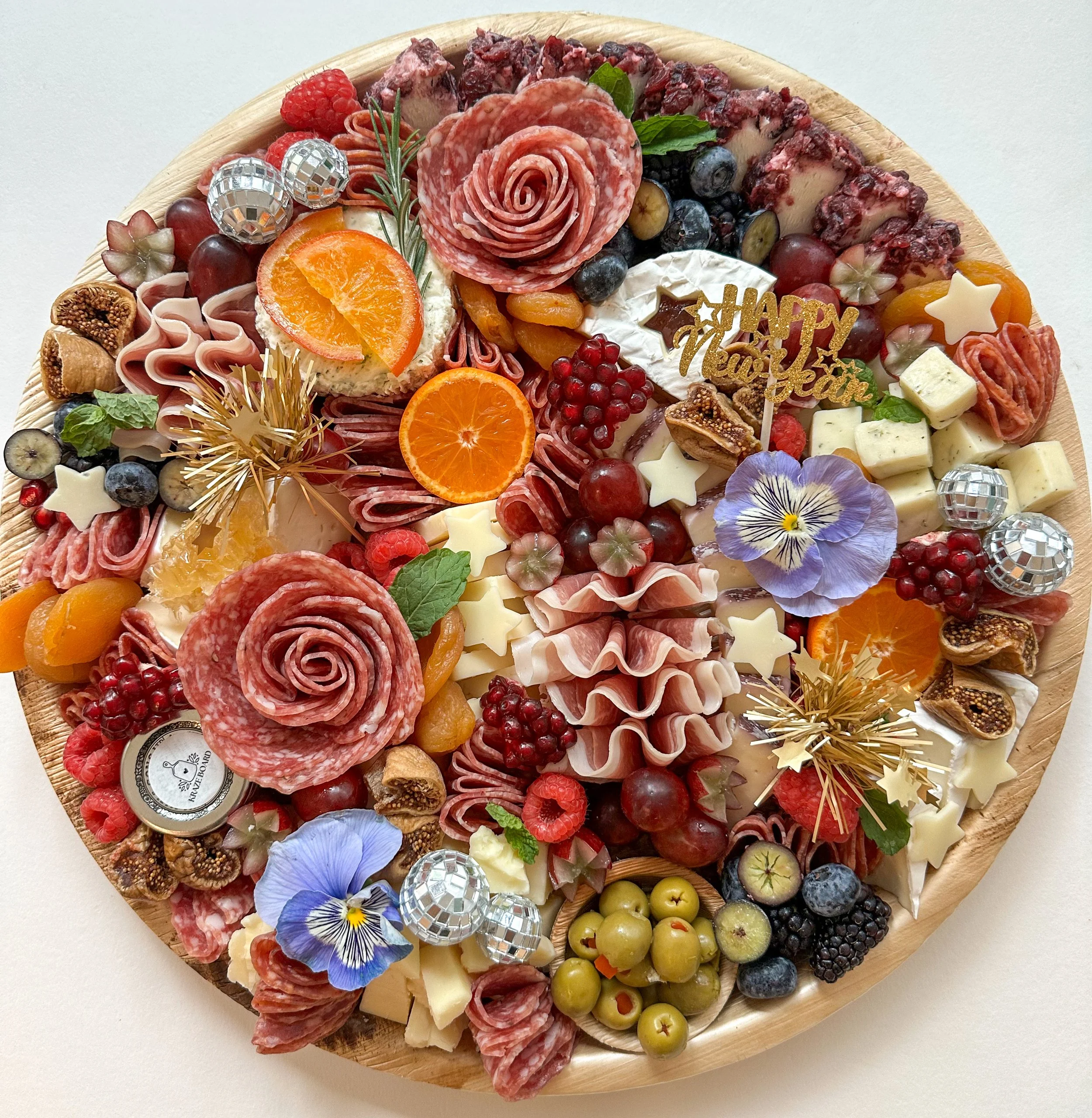 Assorted cheeses, cured meats, fresh fruits, edible flowers, and decorative items on a wooden platter for a celebration. Includes a 