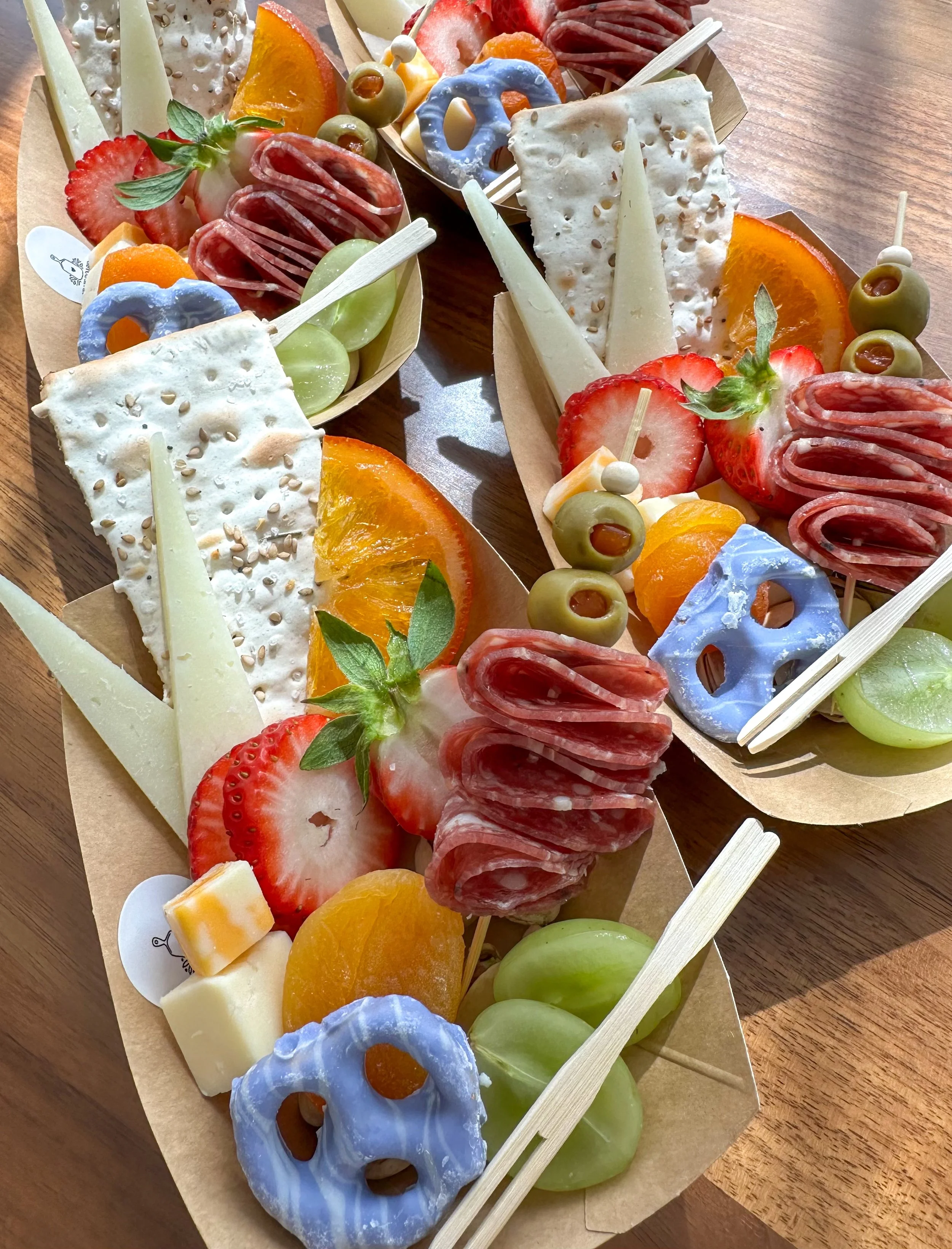 Three wooden trays filled with snacks including strawberries, green grapes, cantaloupe slices, sliced orange, cheese, salami, and colorful pretzels with skewers.
