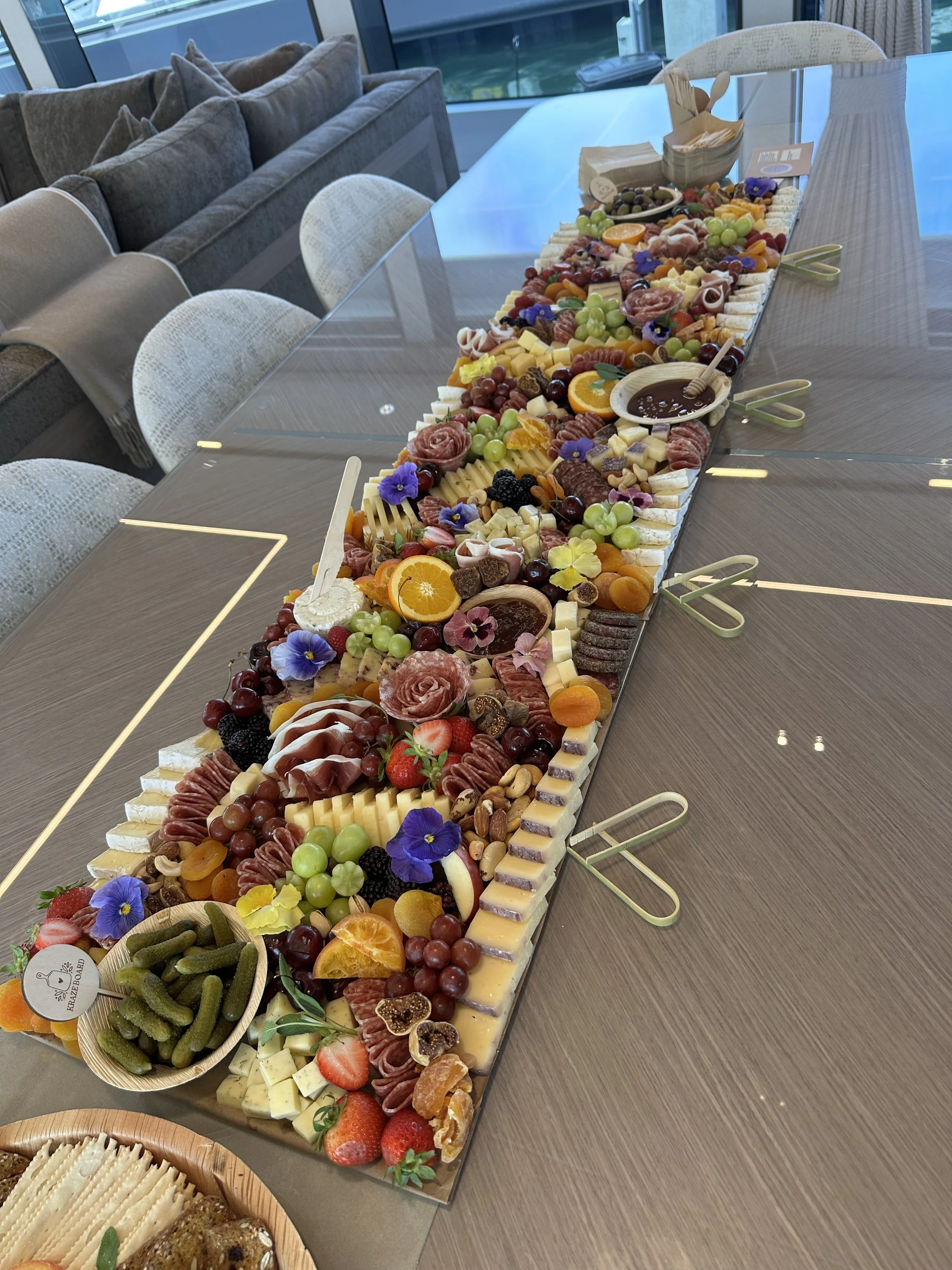 Large cheese and fruit platter on a dining table, featuring assorted cheeses, grapes, strawberries, oranges, blackberries, figs, and edible flowers, decorated for a celebration.