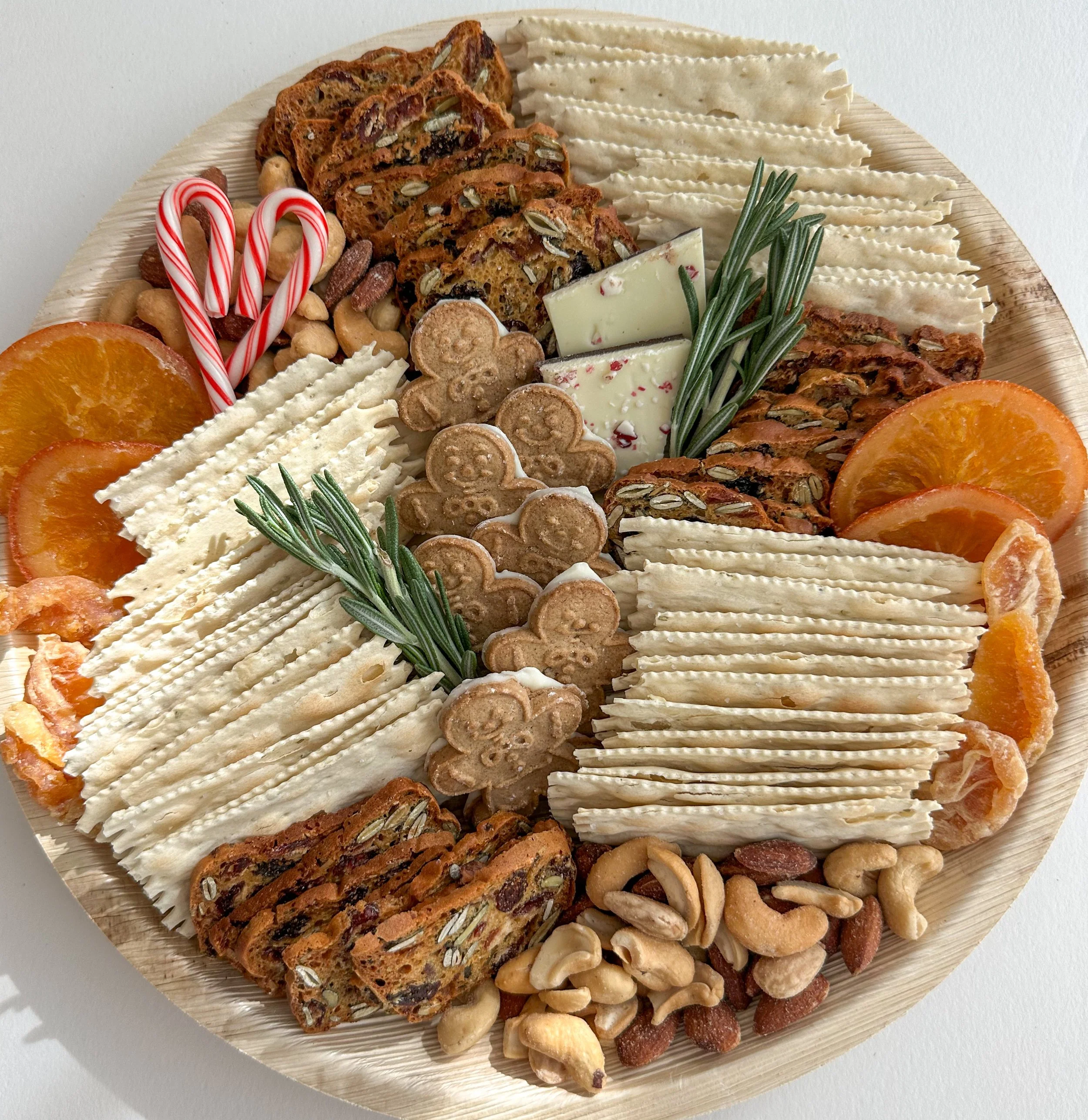 Holiday cheese and snack platter with crackers, dried fruit, nuts, gingerbread cookies, candy canes, white chocolate, and rosemary sprigs on a round wooden tray.