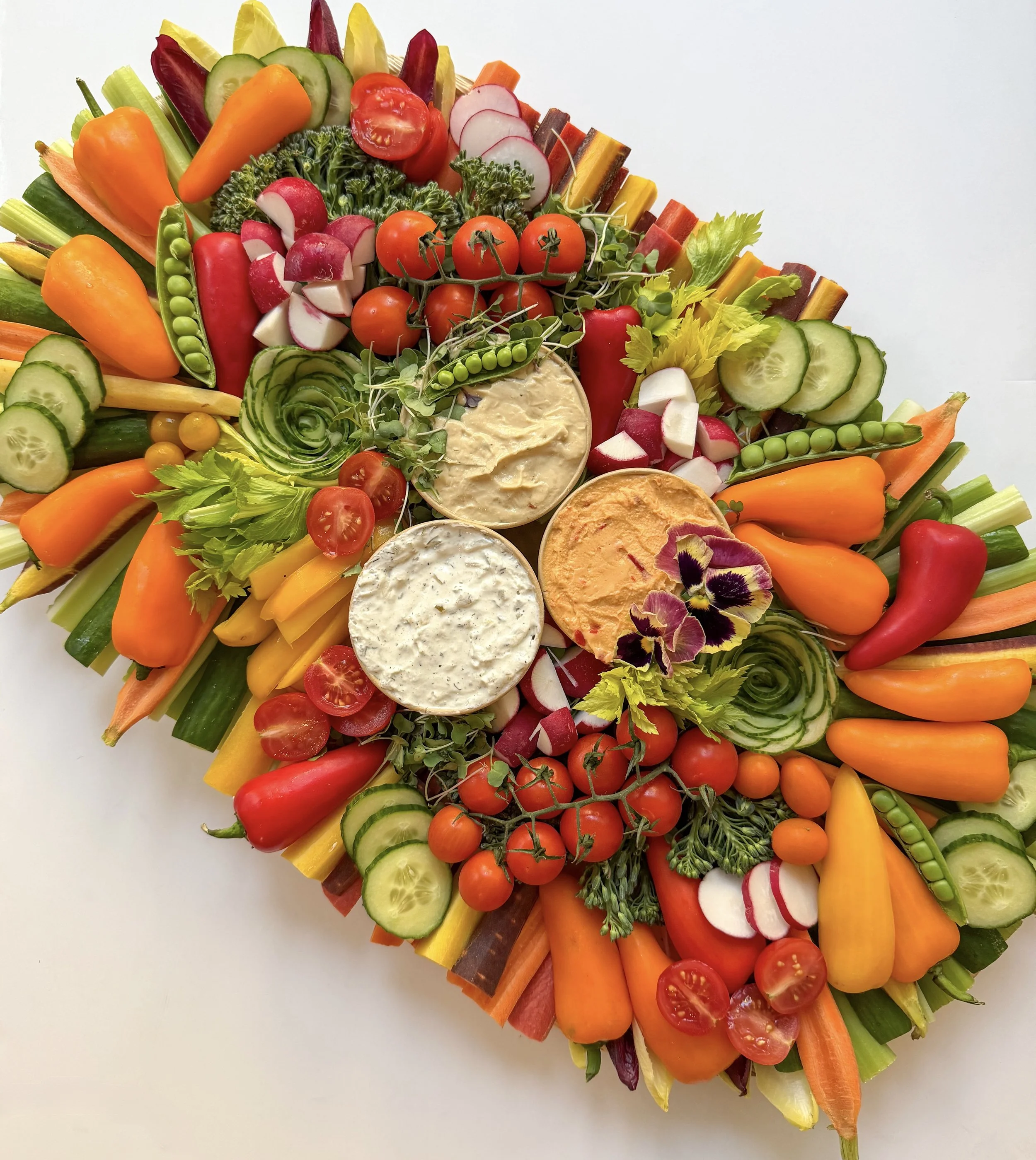 Colorful vegetable platter with cherry tomatoes, cucumber slices, radish slices, bell peppers, cauliflower, broccoli, and two bowls of creamy dips decorated with edible flowers.