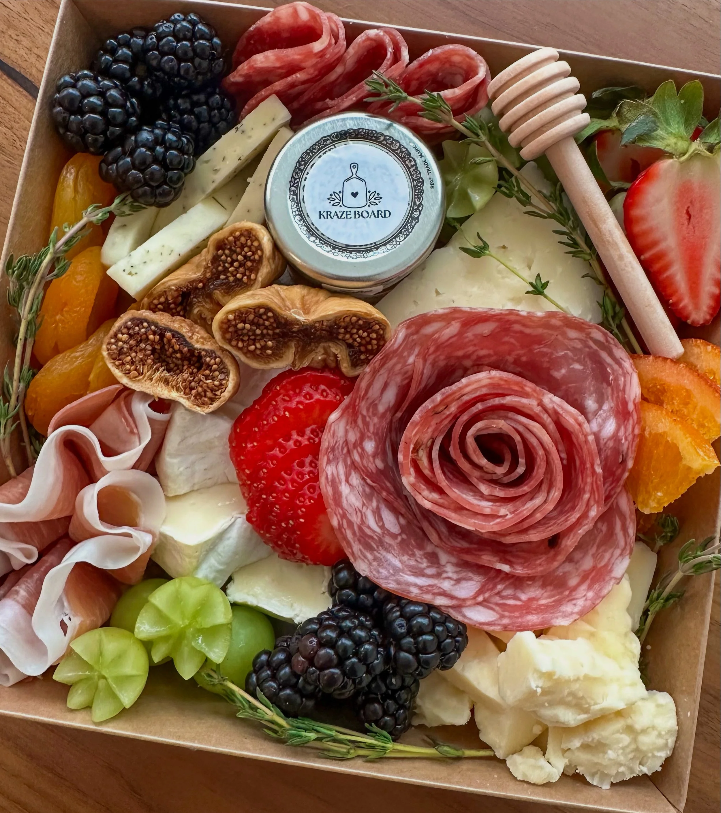 A charcuterie board with various cheeses, sliced meats, fresh strawberries, blackberries, green grapes, figs, figs, and slices of cucumber and honey. There is also a honey dipper, herbs, and a small jar labeled 'Kraze Board' in the center.