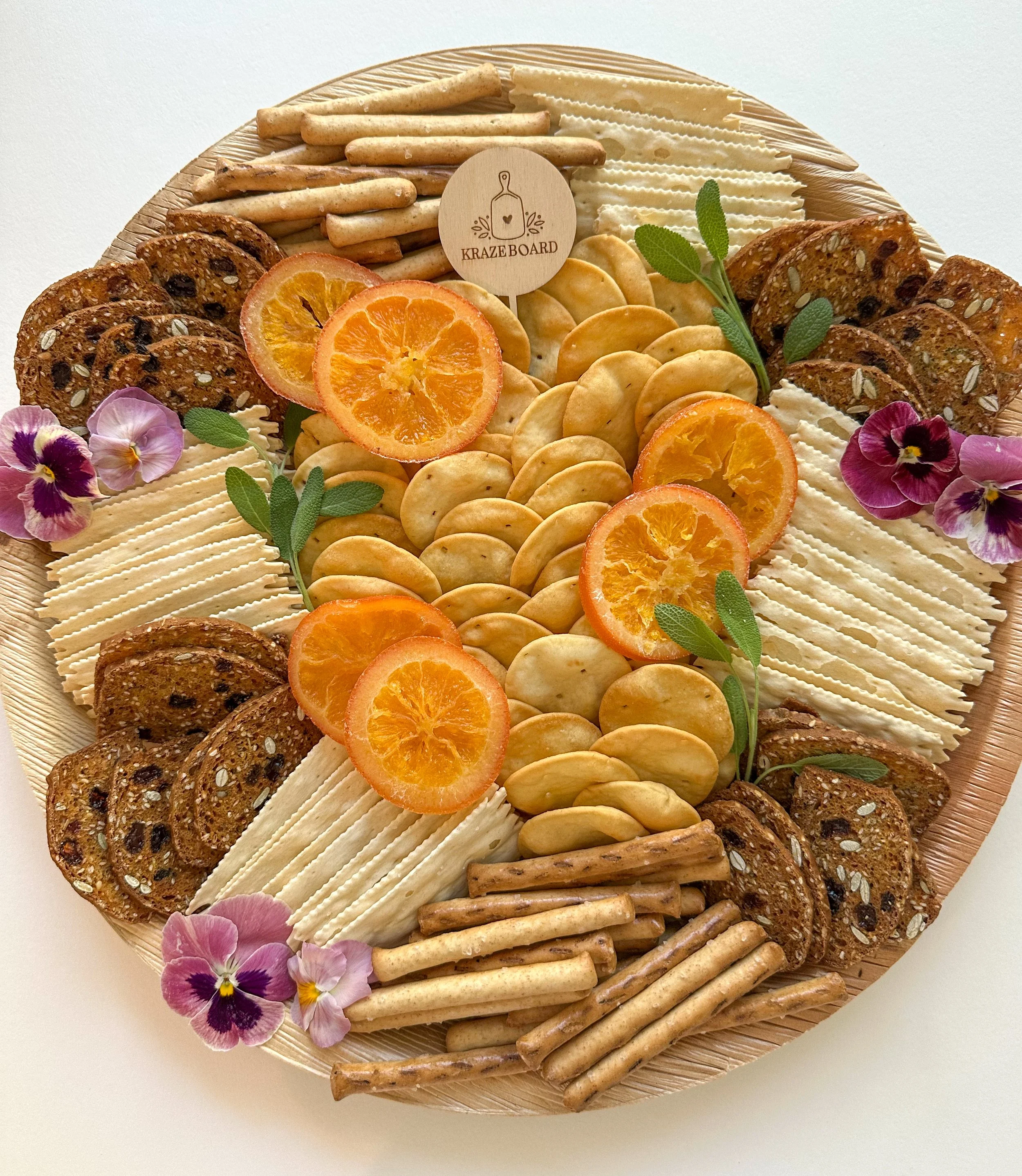 Cheese and cracker platter with orange slices, edible flowers, and greenery, arranged on a round eco-friendly palm leaf disposable board.