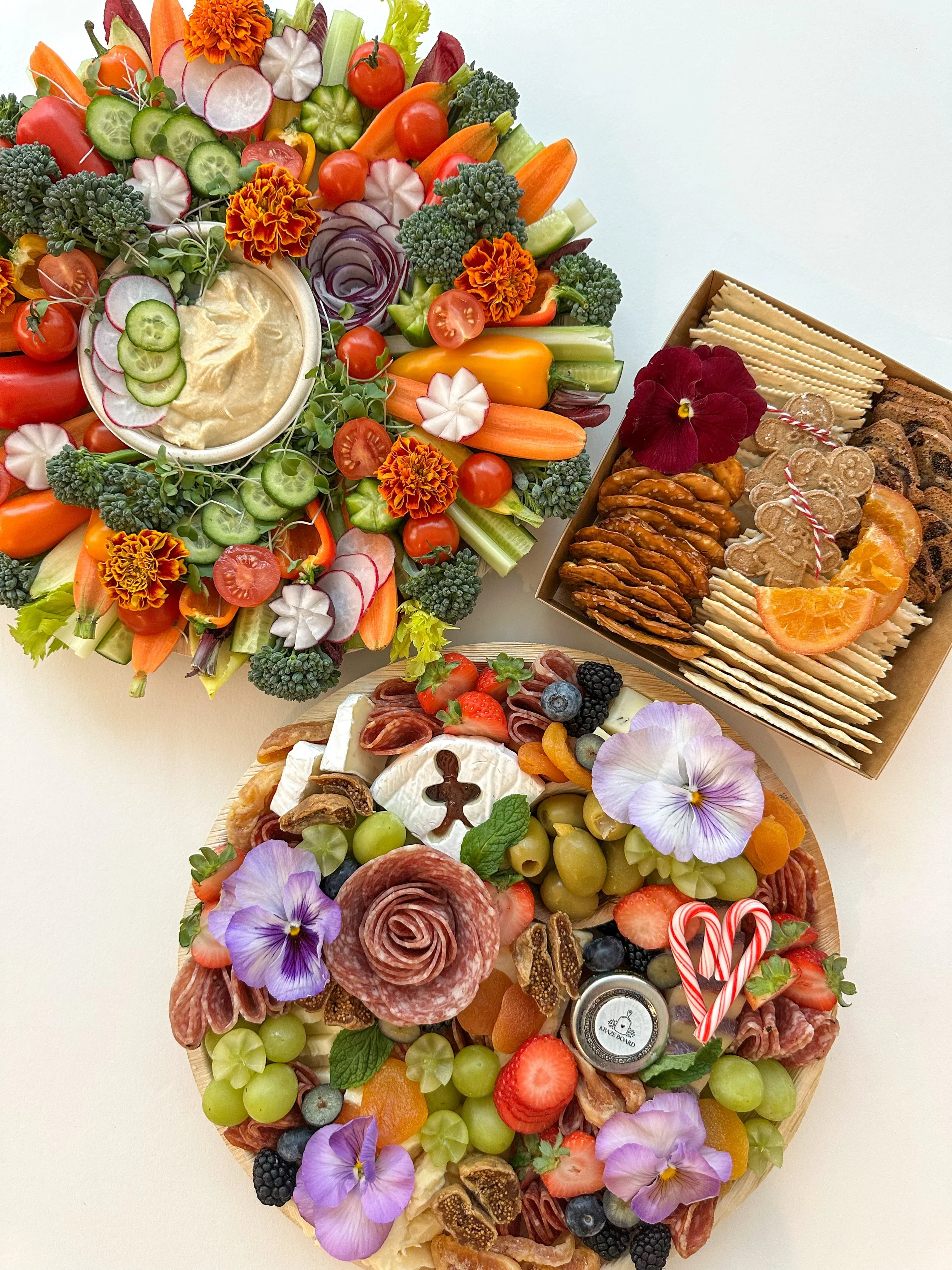 A colorful charcuterie and cheese platter with grapes, strawberries, berries, sliced vegetables, edible flowers, nuts, and assorted cheeses, alongside a bowl of hummus, and a box of assorted cookies and crackers.