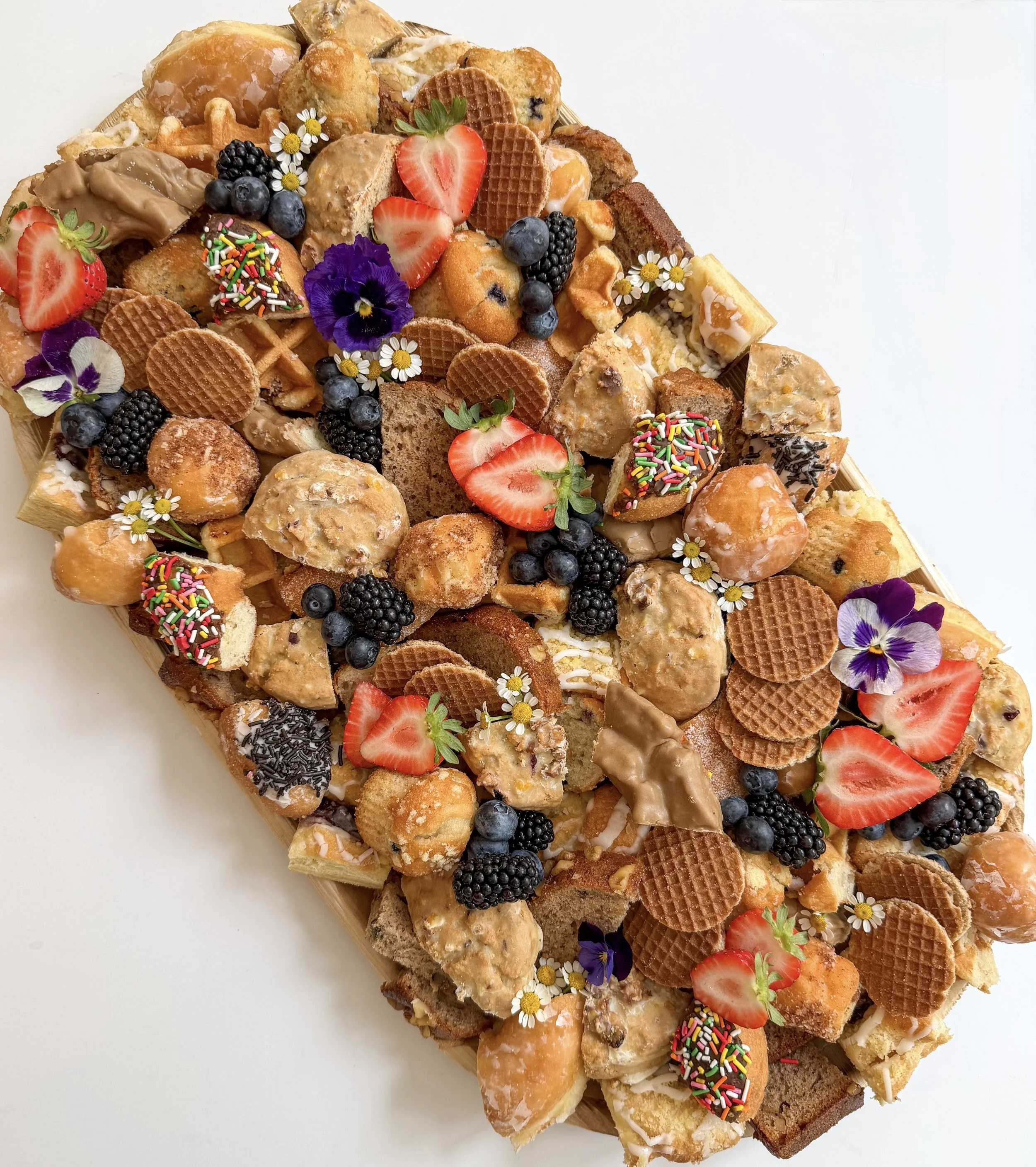 A large fruit and dessert platter with various fruits, cookies, pastries, edible flowers, and a donut with sprinkles.