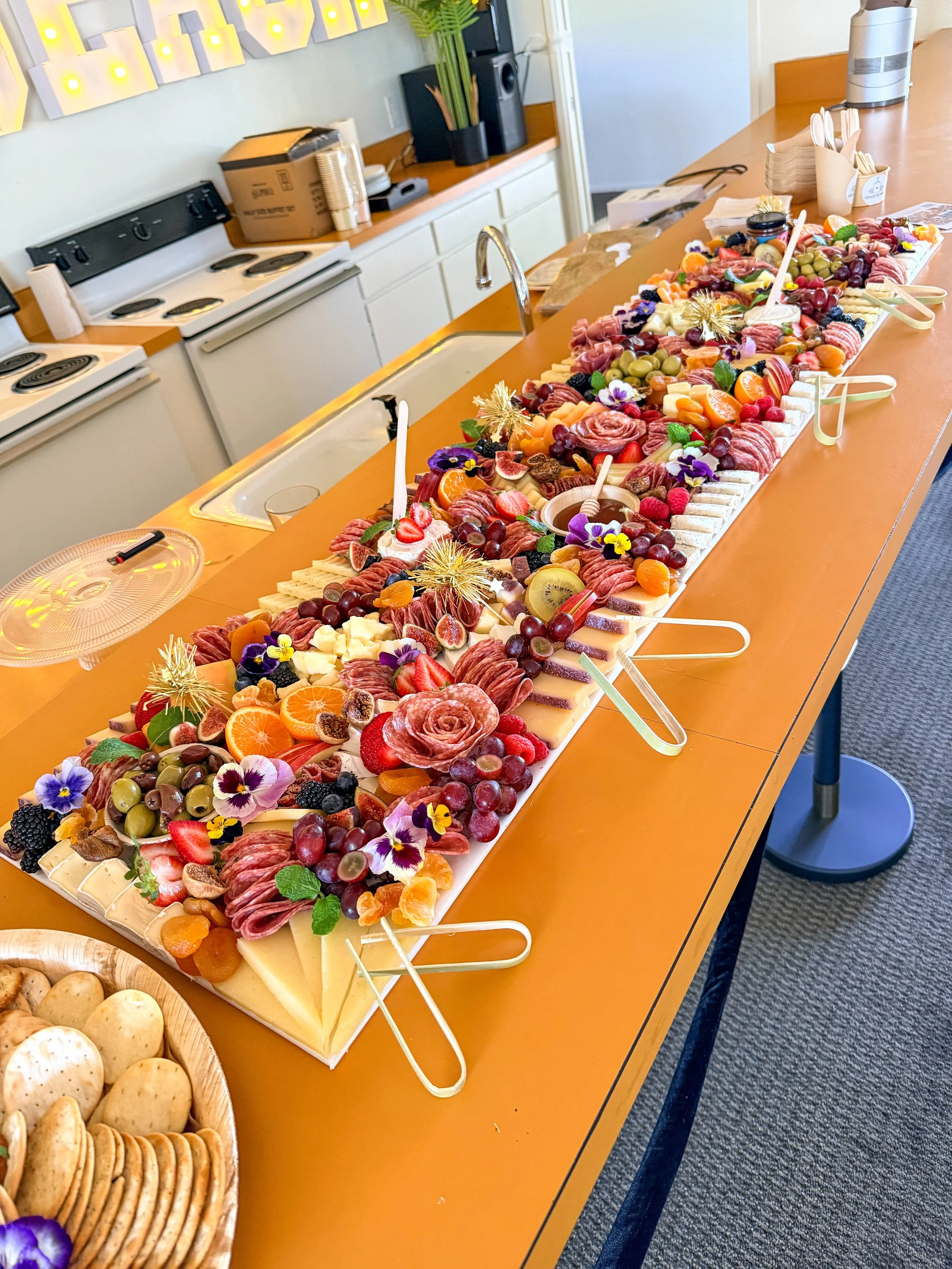 A long buffet table with an assortment of cheeses, fruits, berries, and garnishes, decorated with edible flowers, set up for a gathering or event in a room with kitchen appliances and a cake on a tray.